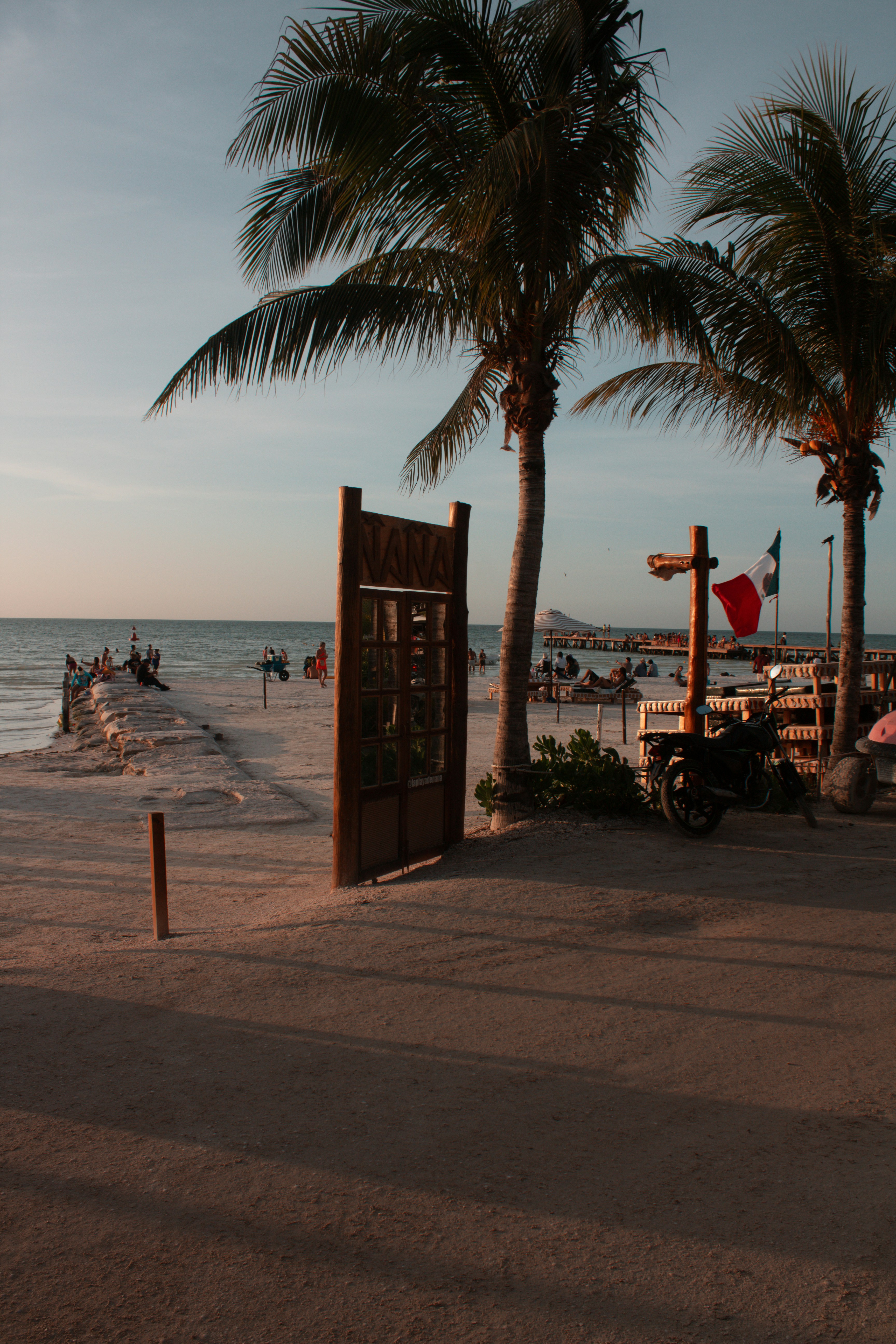A wooden door frame on a sandy beach with palm trees and a Mexican flag