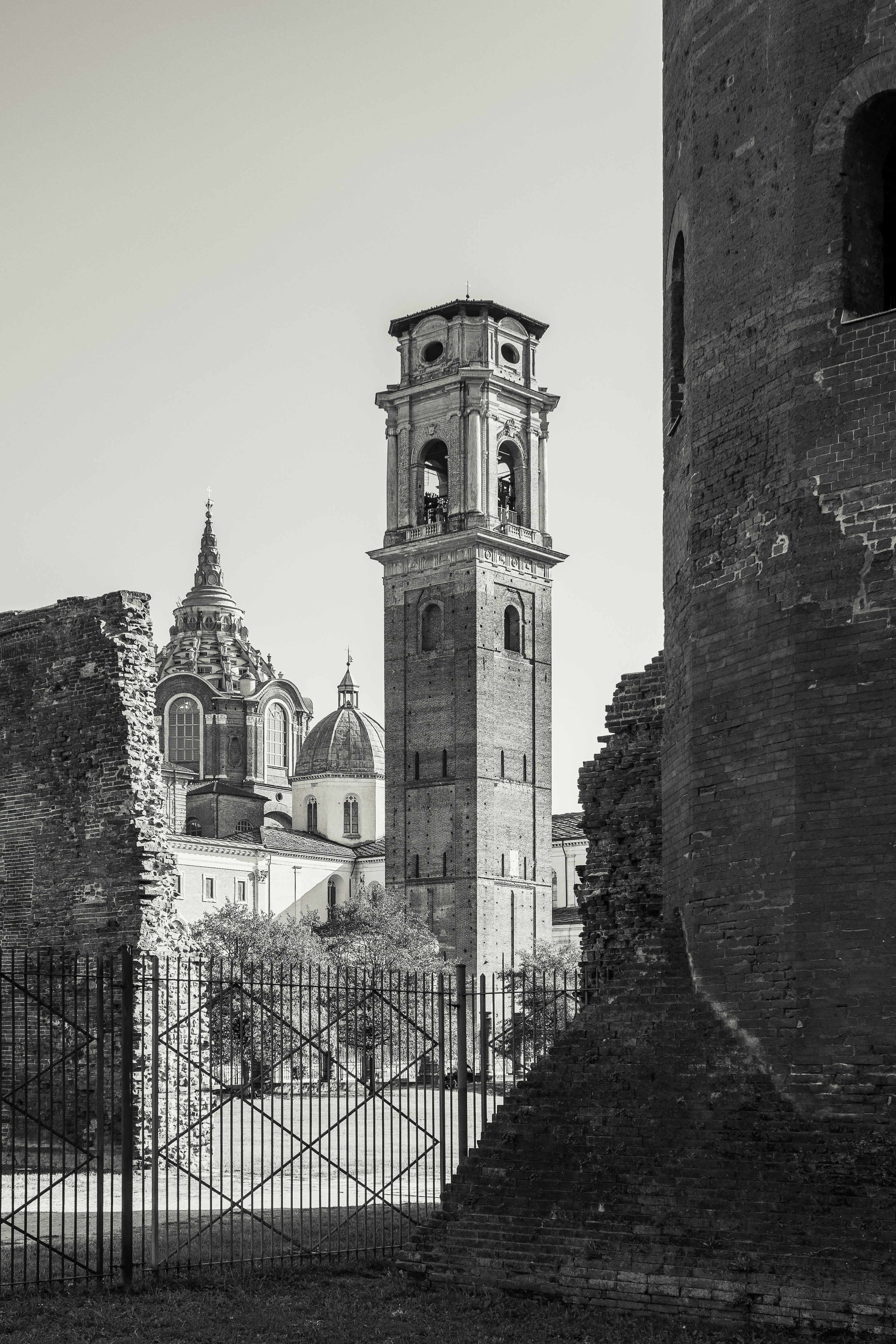 A tall brick bell tower and church domes behind a metal fence in Turin