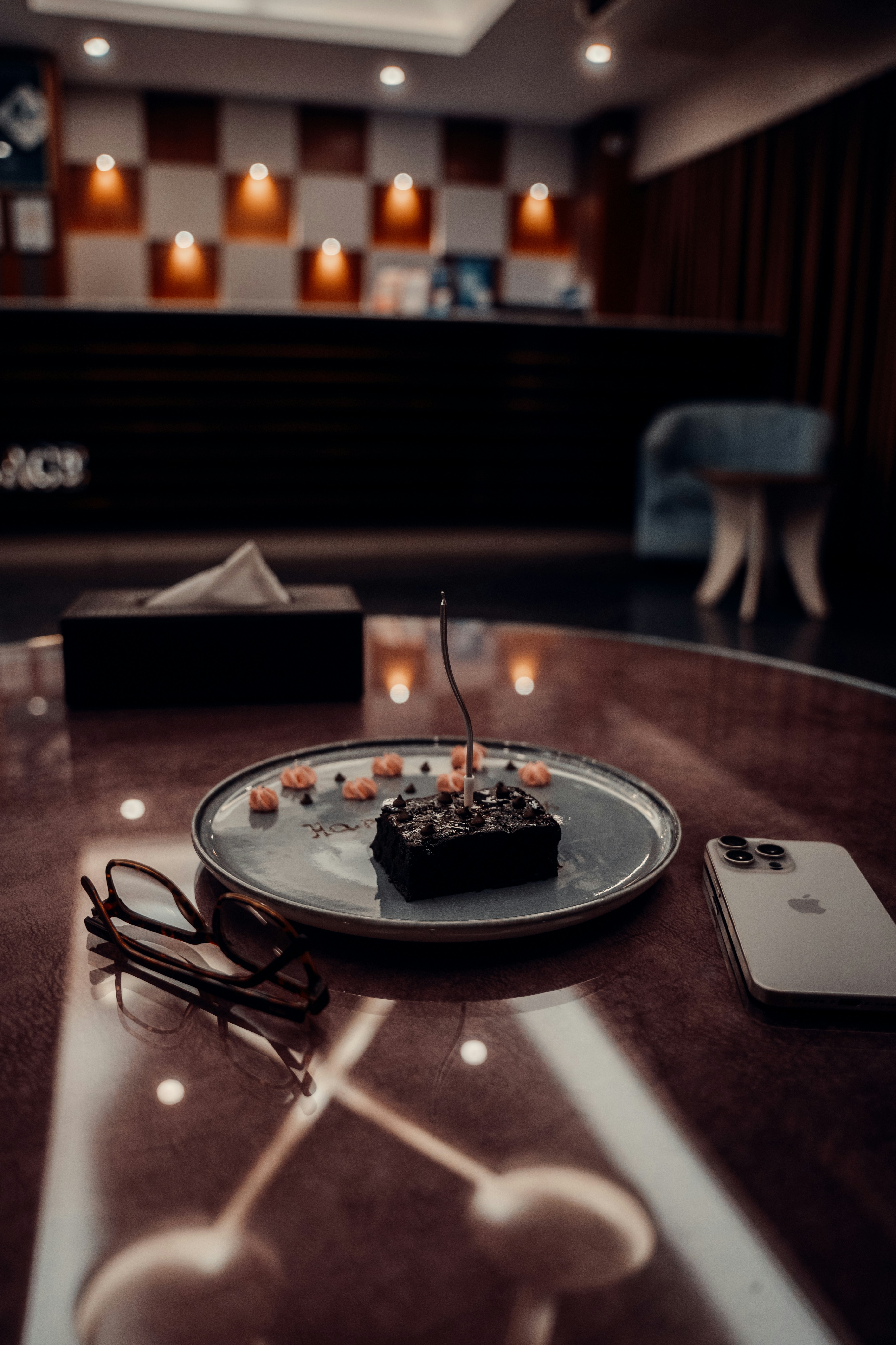 A chocolate brownie with a single candle on a plate beside glasses and phone