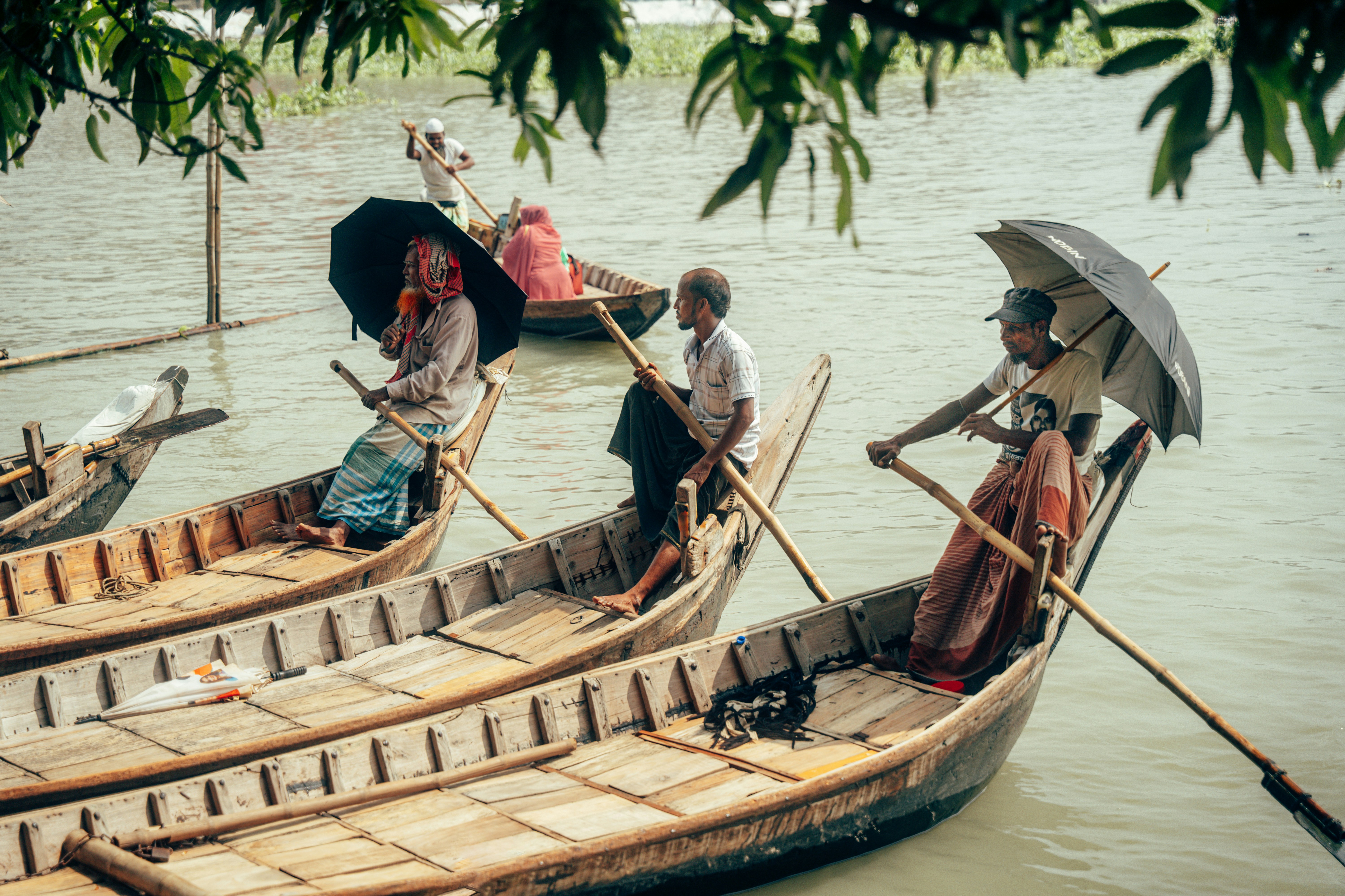 People rowing wooden boats on a river, some holding umbrellas for shade