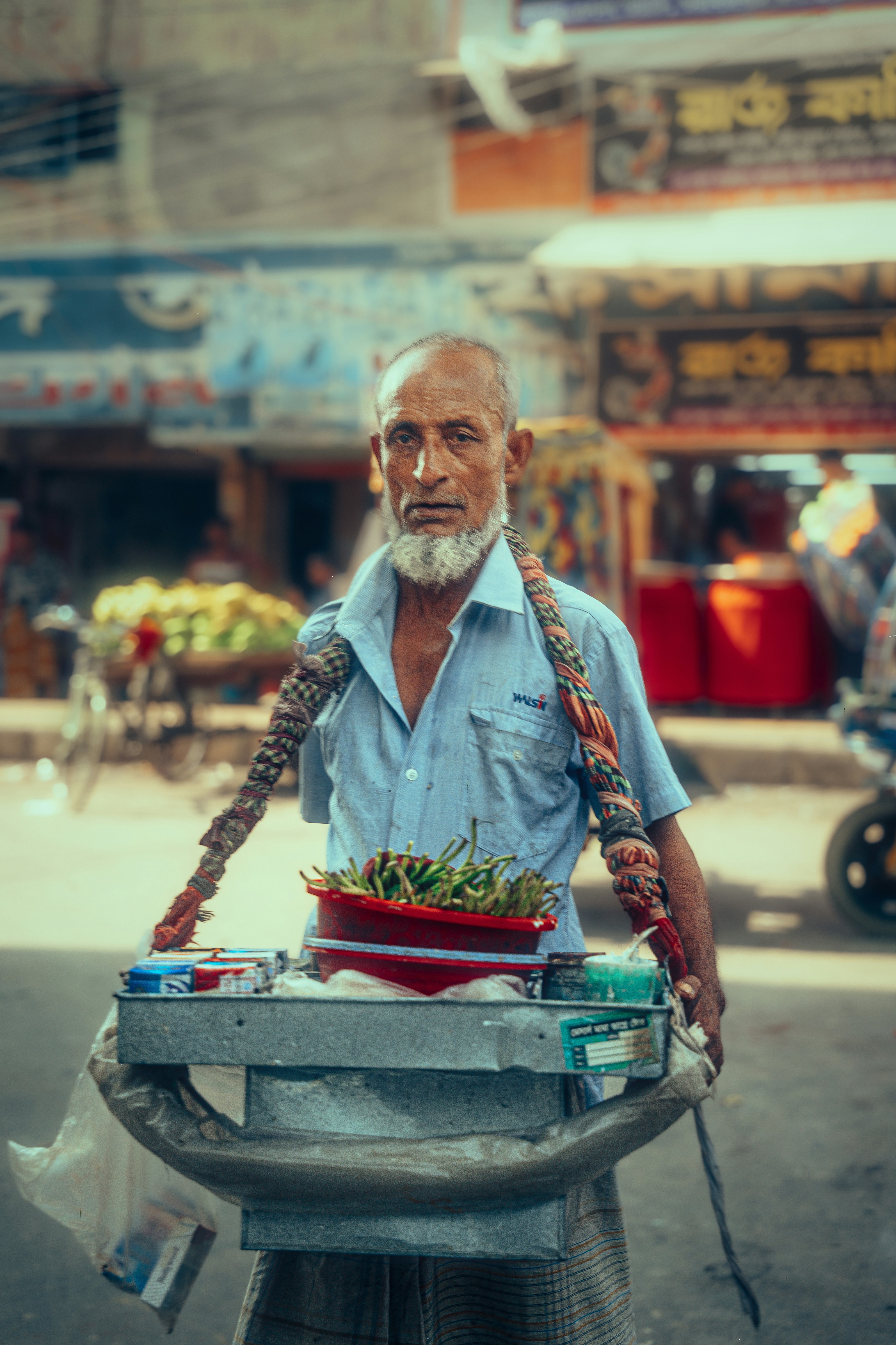An elderly man with a white beard carrying a metal tray of green chilies