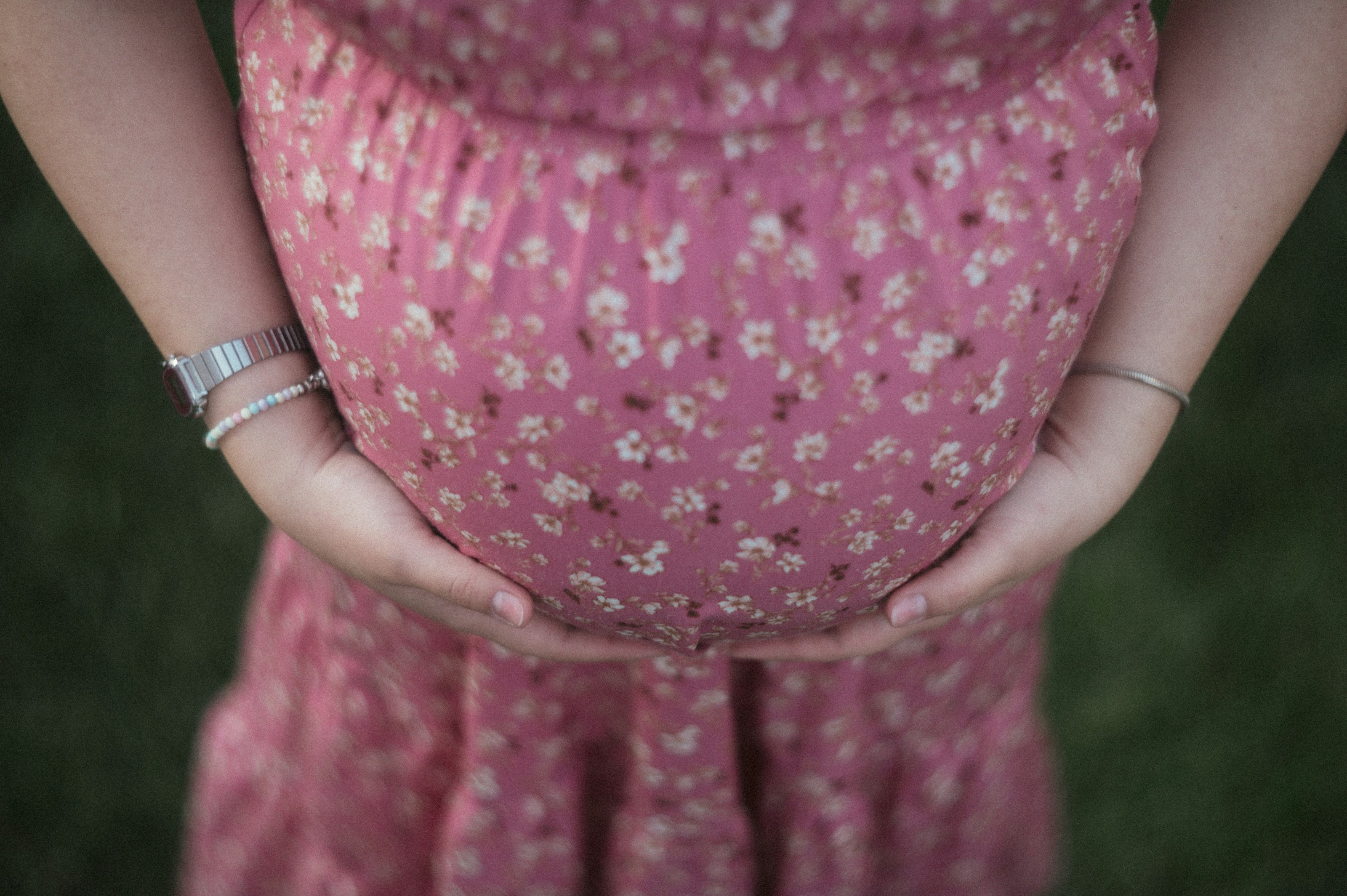 A person in a floral pink dress cradling their pregnant belly with both hands
