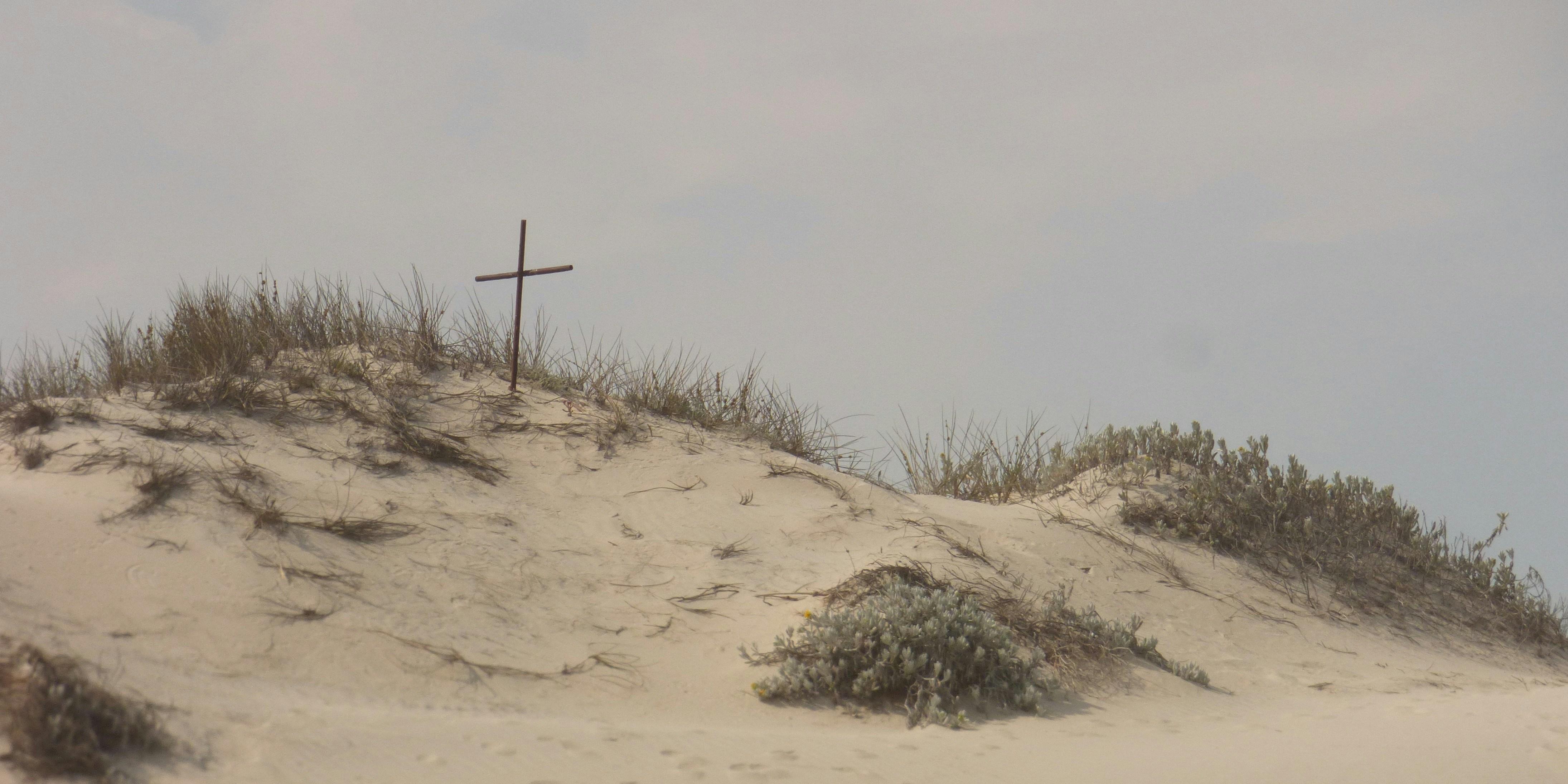 A thin metal cross on a sandy dune with sparse grass