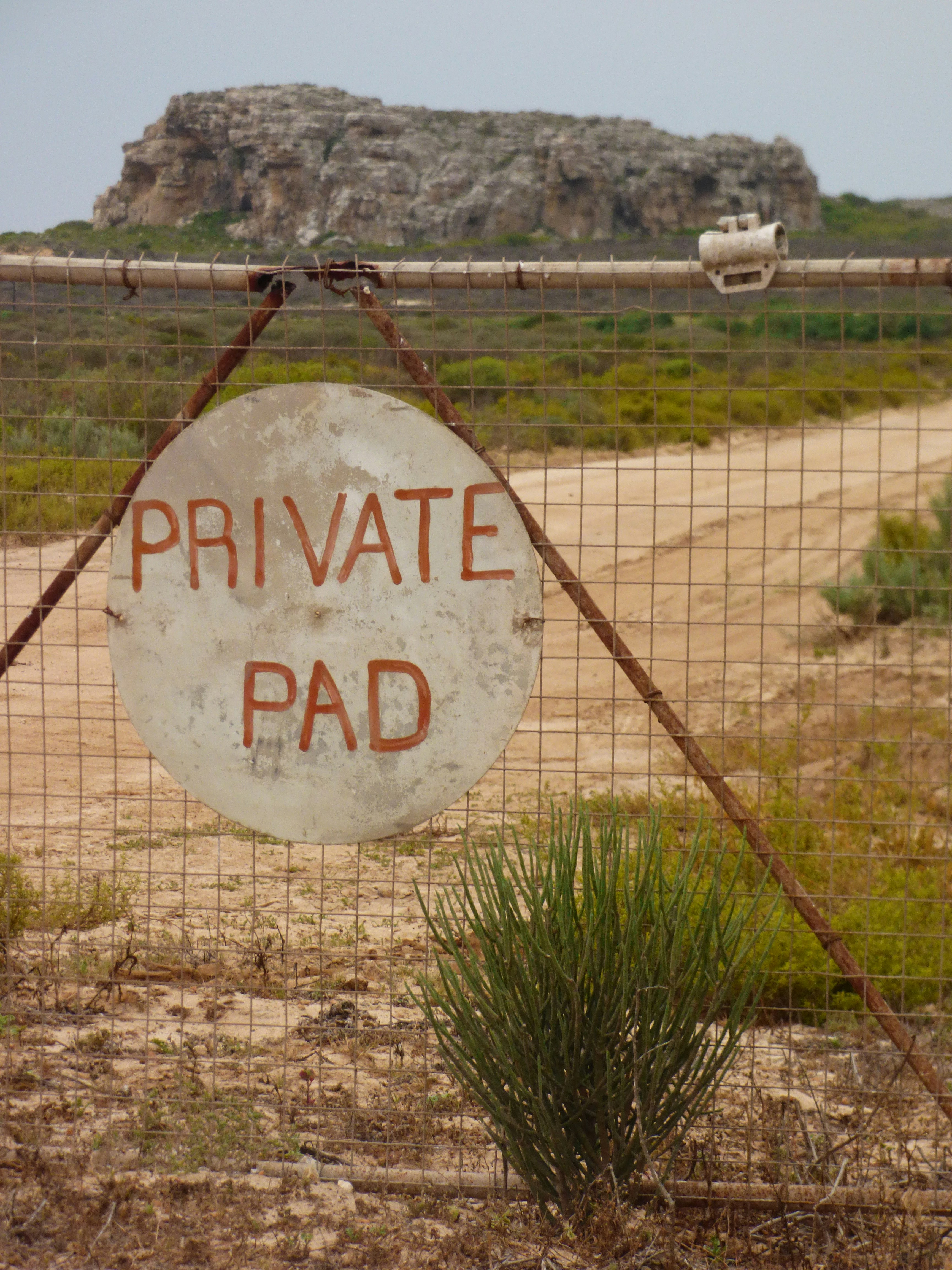 Uma placa redonda com a inscrição "PRIVATE PAD" em uma cerca de arame antes de uma estrada de terra