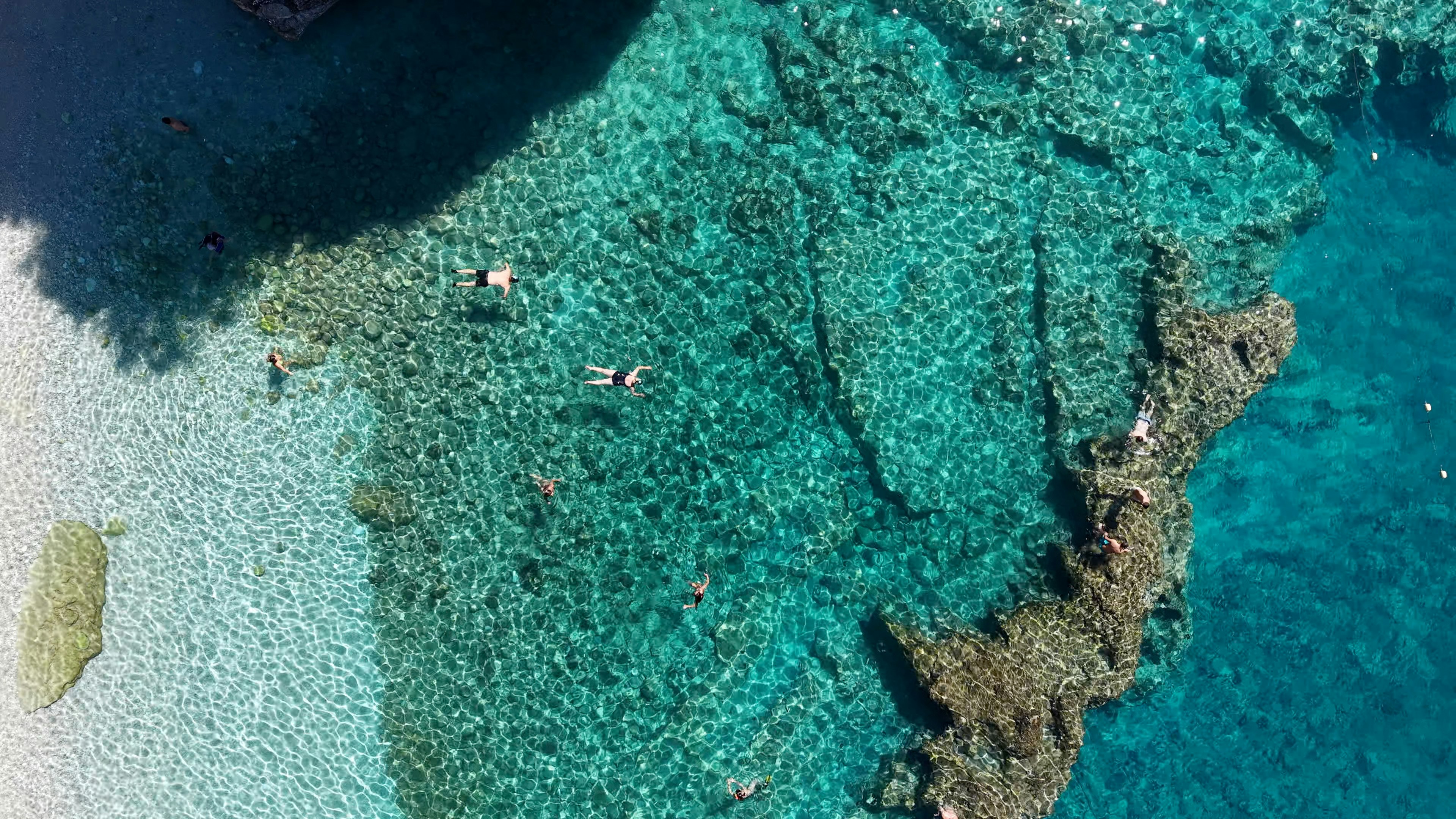 People swimming in clear turquoise water near a rocky coastline from a drone view
