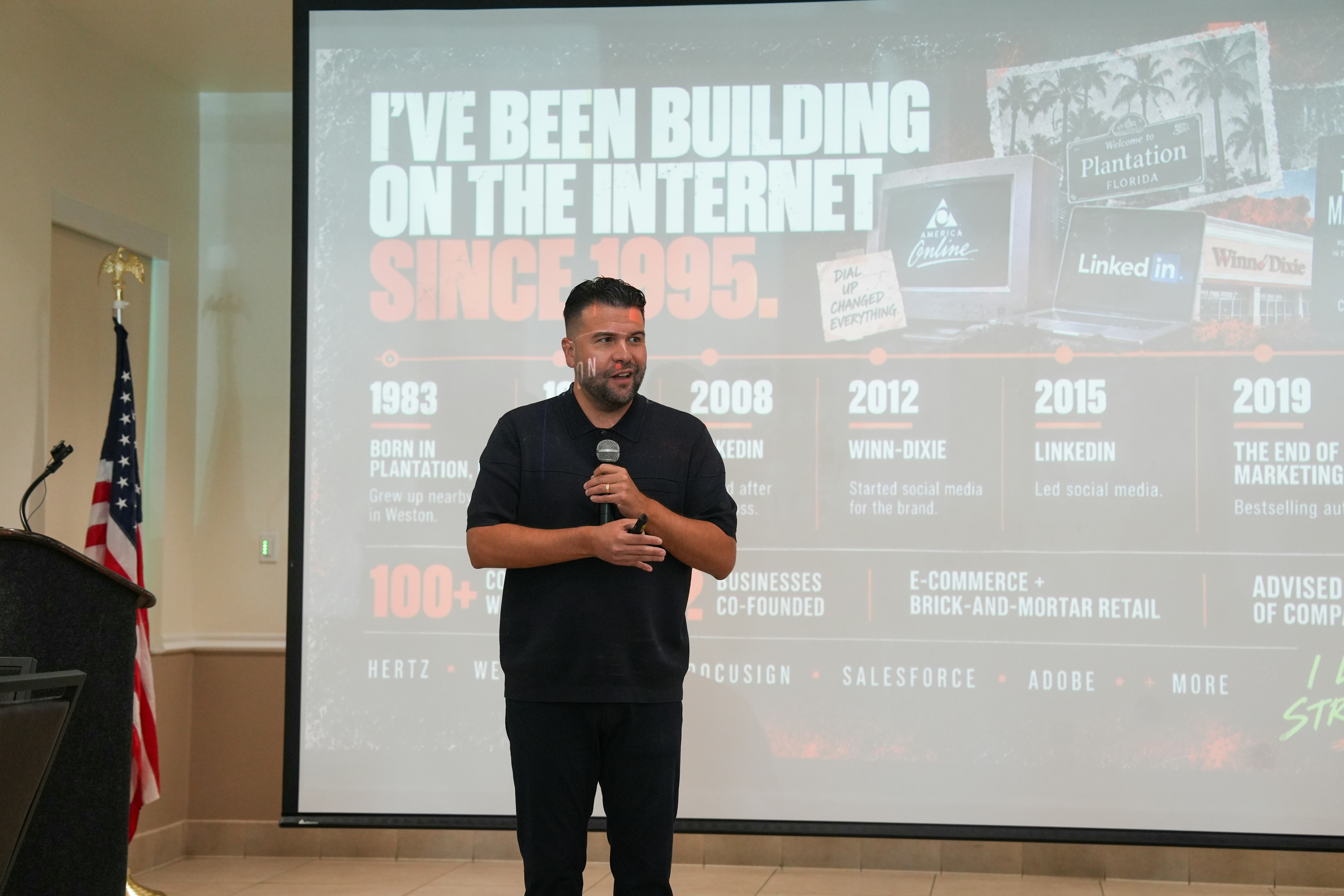 A man in a black polo shirt speaking into a microphone before a presentation
