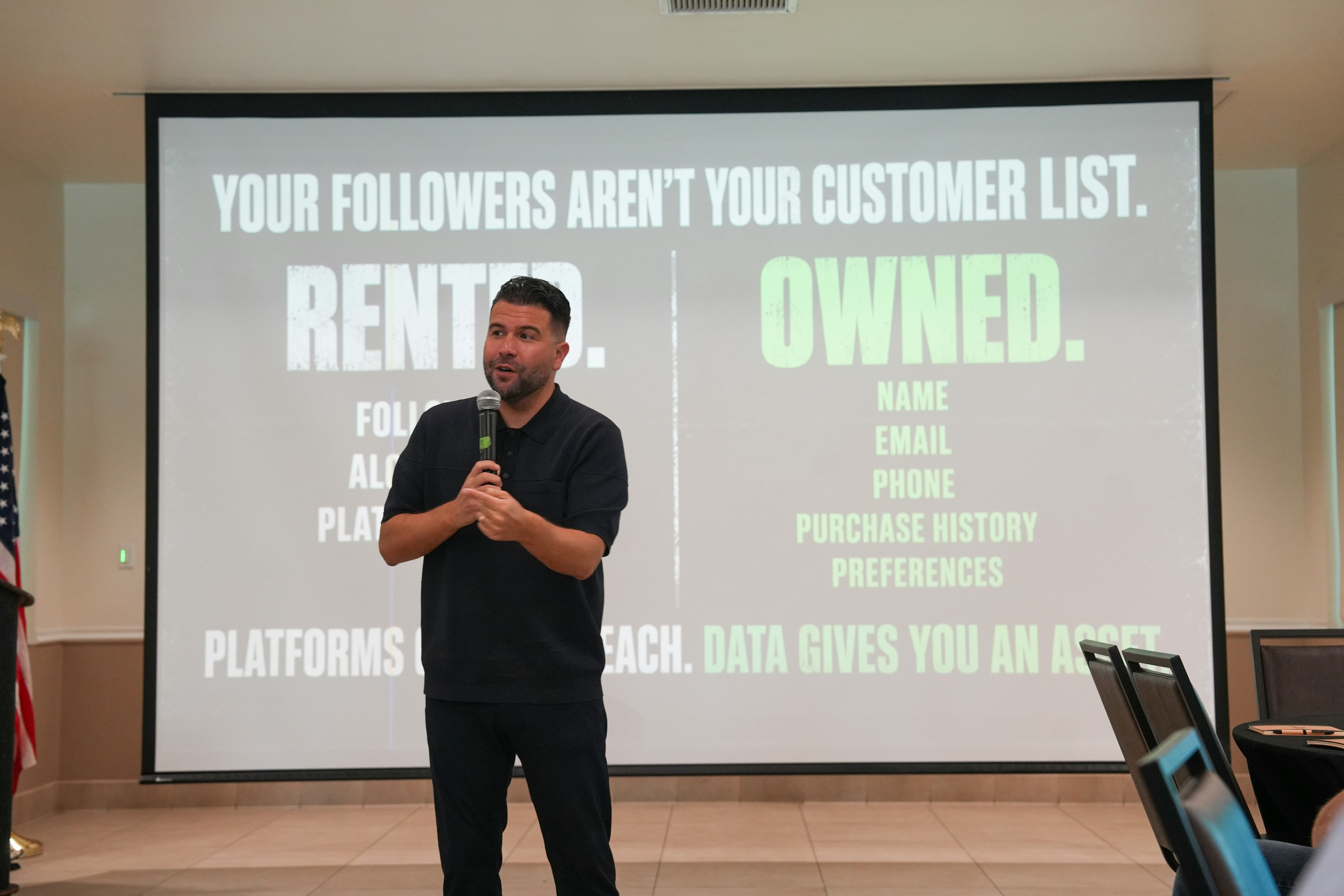 A man in a black polo shirt speaking into a microphone before a presentation