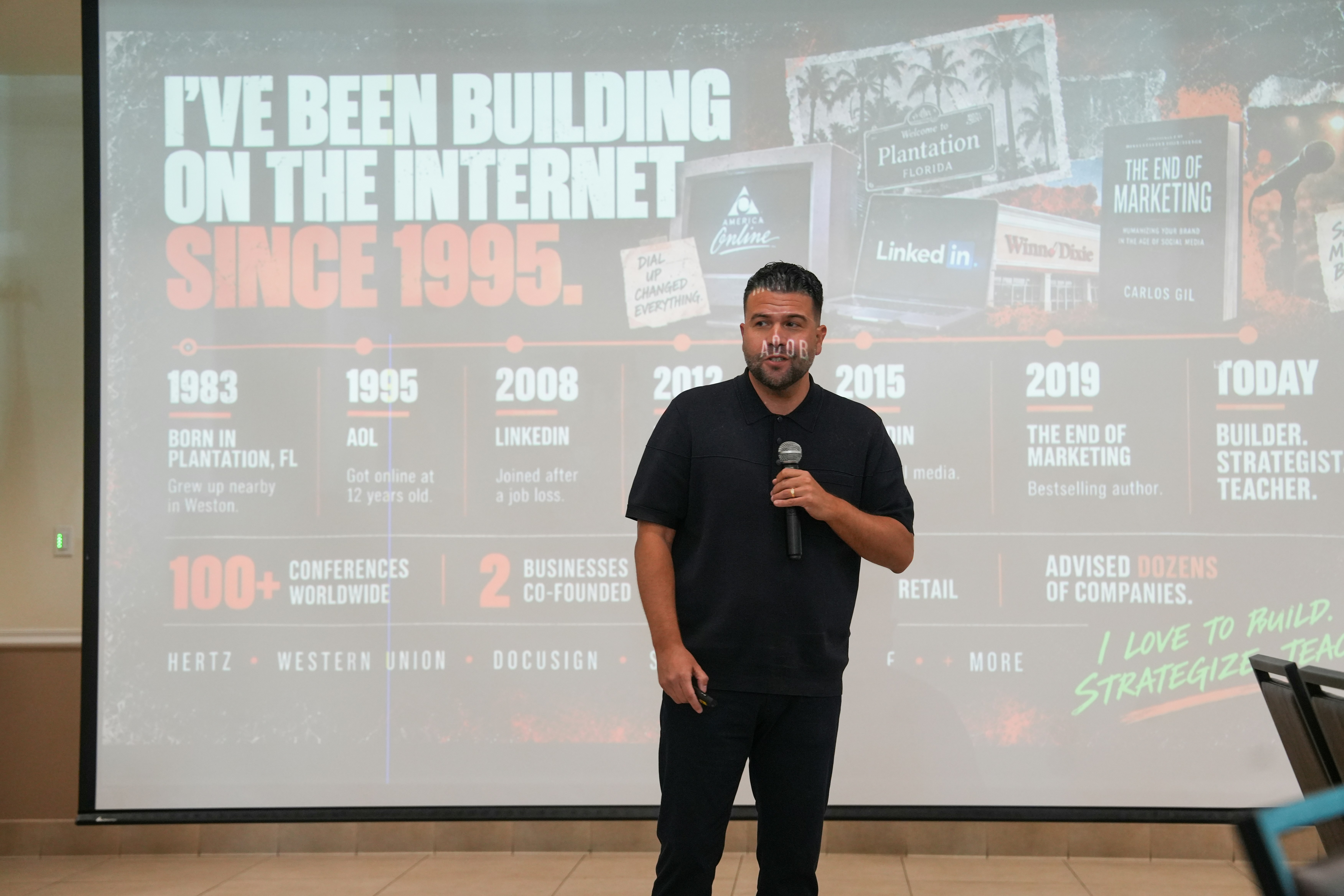 A man in a black polo shirt speaking into a microphone before a presentation