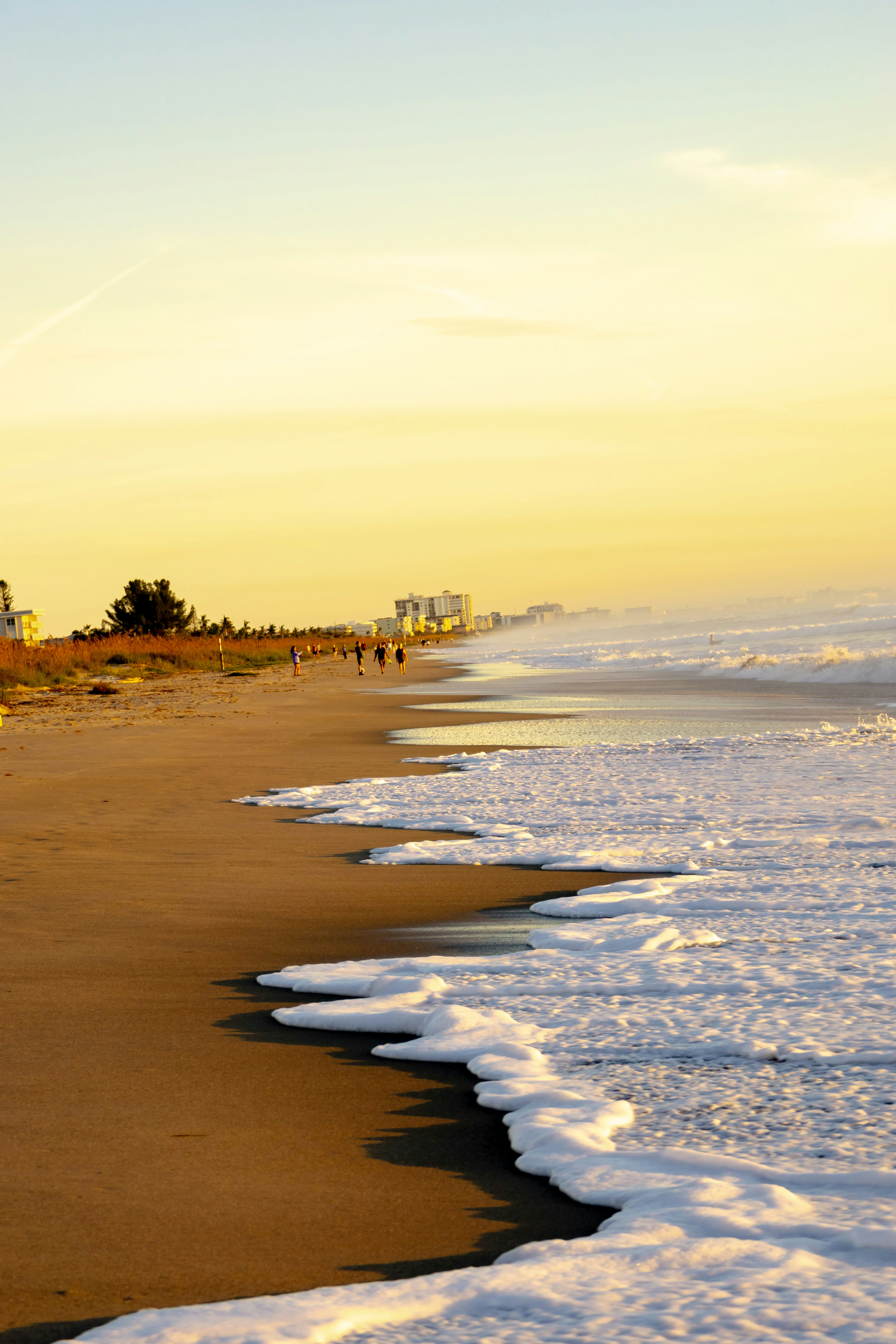 People walking along a sandy beach with white sea foam under golden light