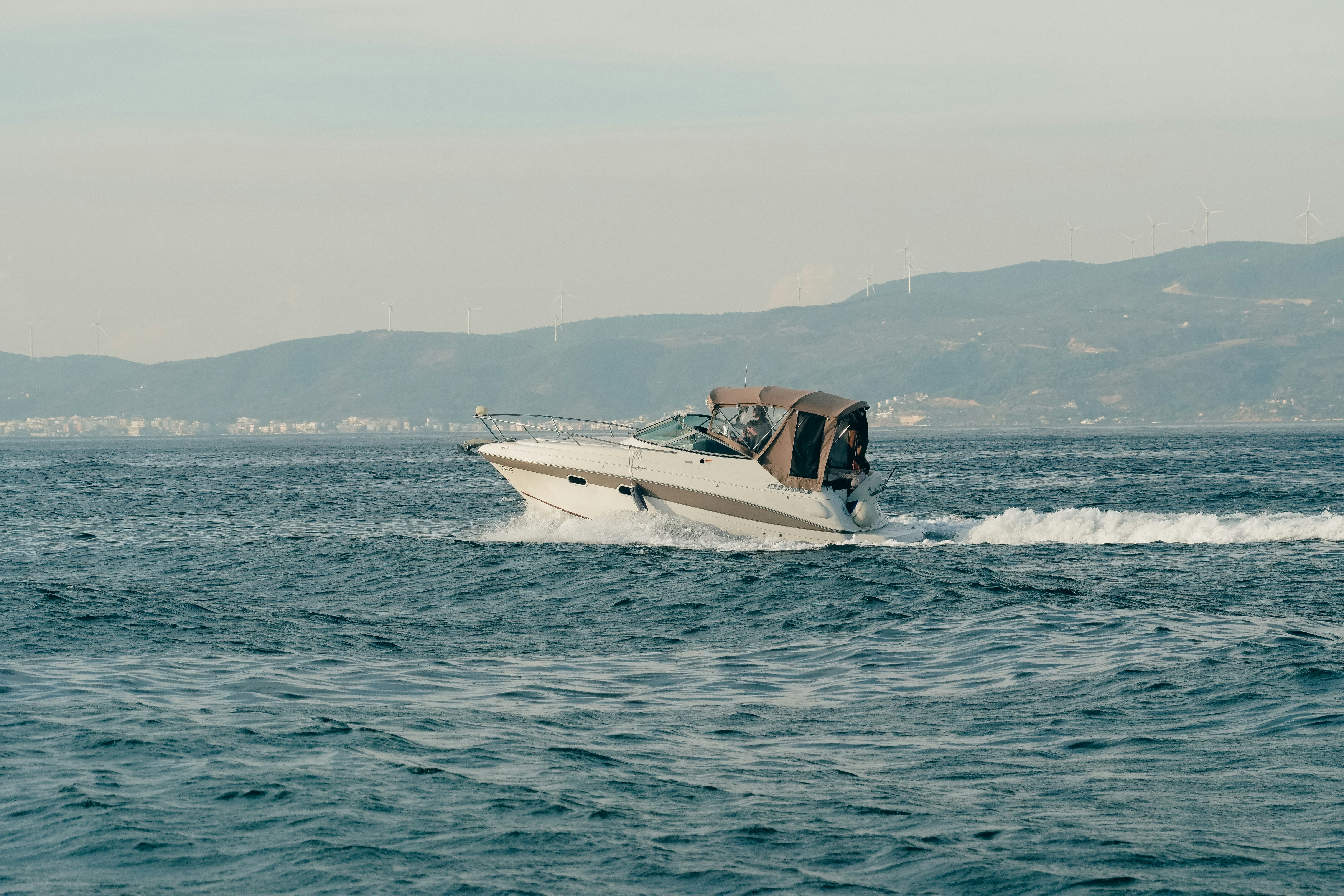 A white motorboat moving across choppy water with distant hills and wind turbines