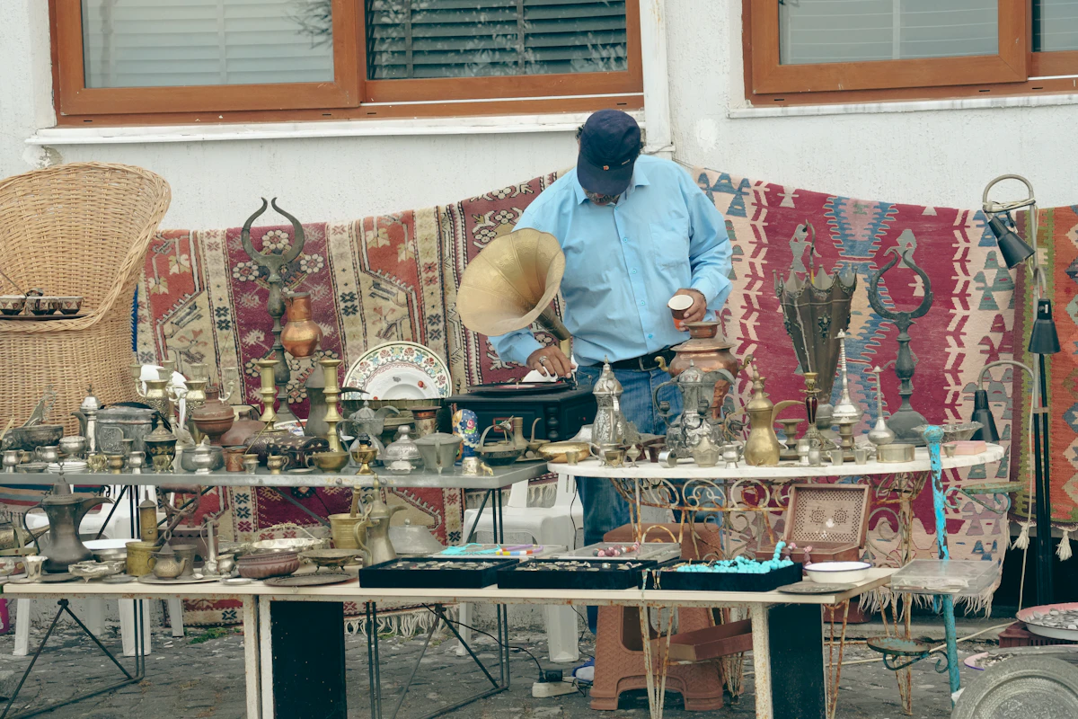 Un homme en chemise bleu clair et casquette manipule un gramophone à pavillon doré derrière un étal en plein air couvert d'objets en cuivre, devant des tapis tendus contre un mur
