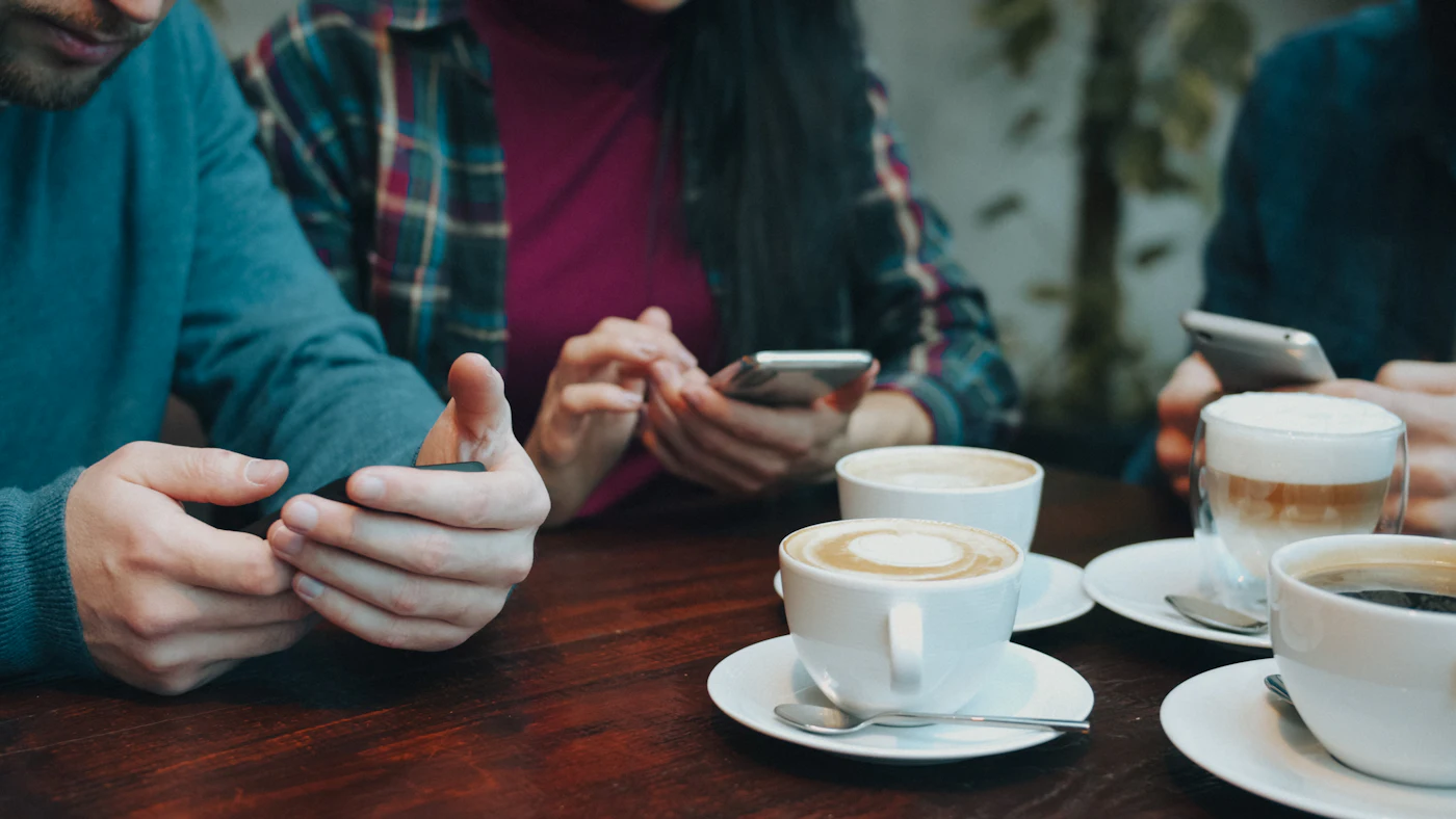 Three people at a wooden table with coffee, looking at their phones.