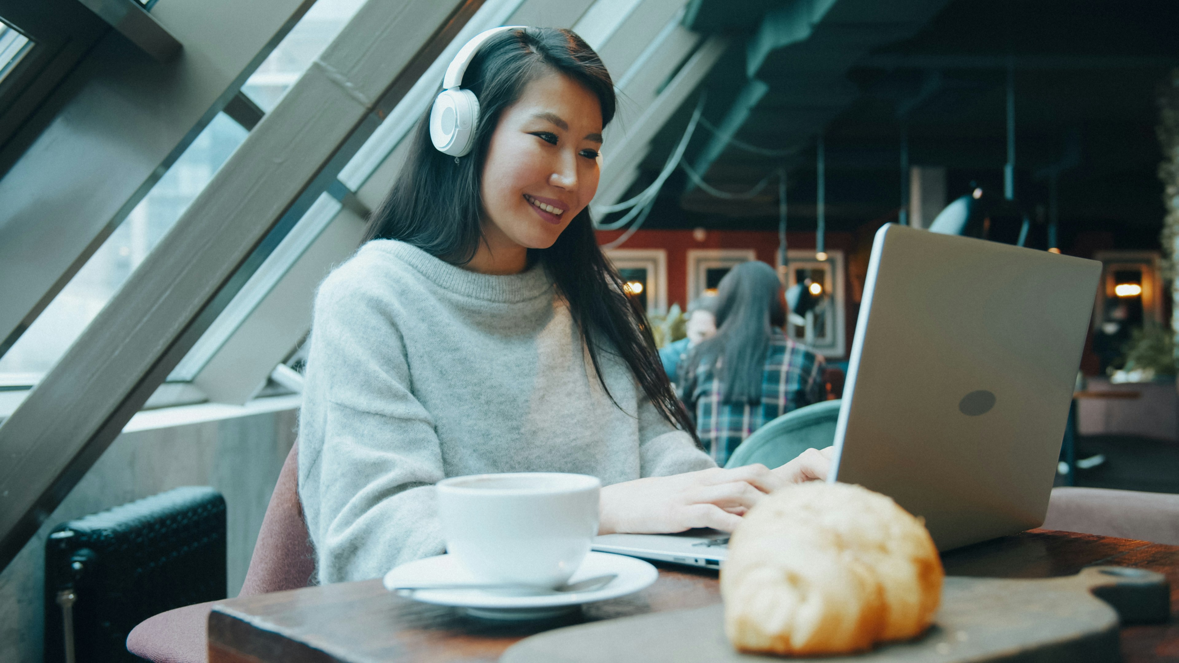 A woman wearing white headphones using a laptop in a cafe with a croissant