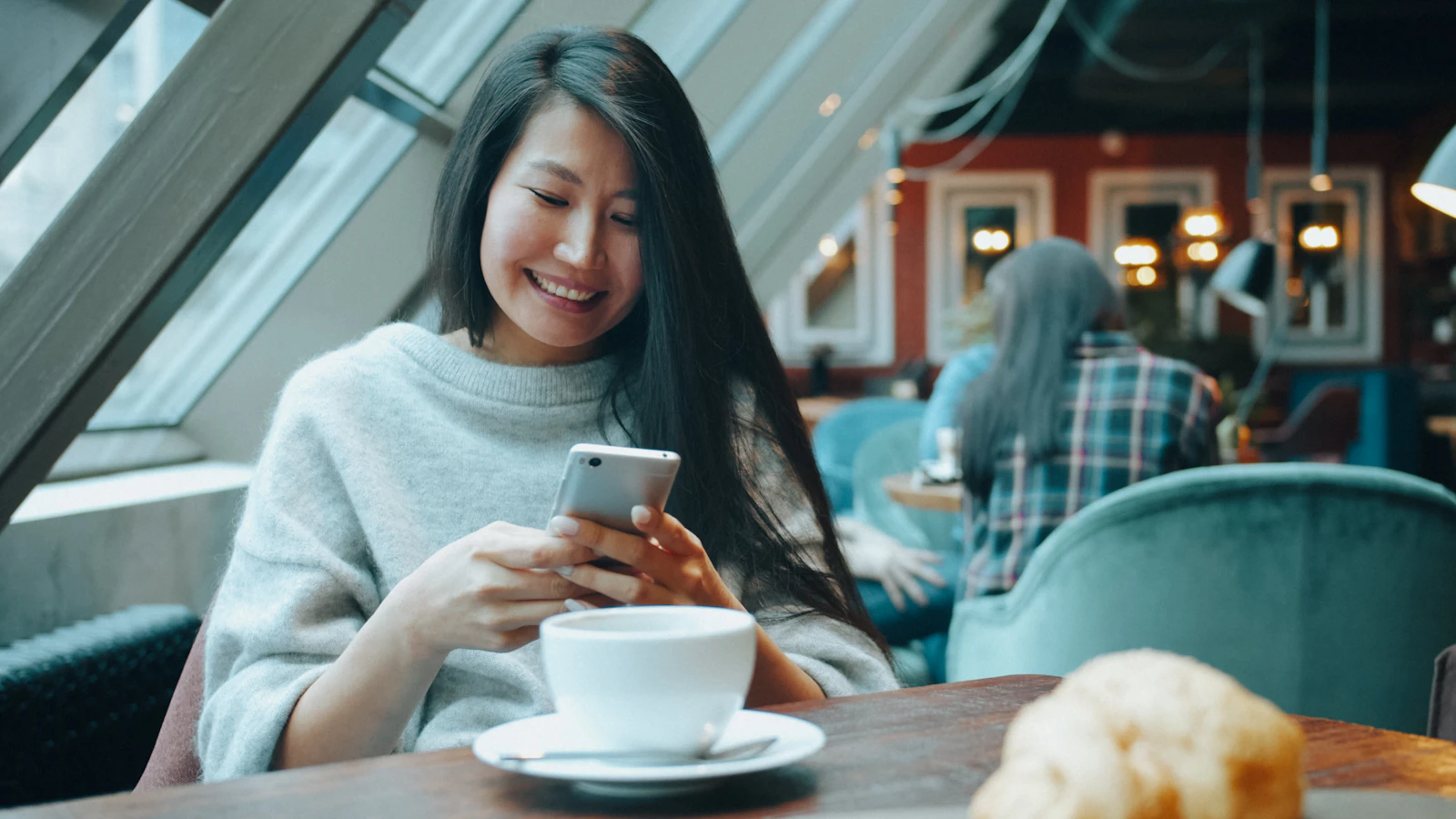 A smiling woman in a grey sweater using a smartphone at a cafe table