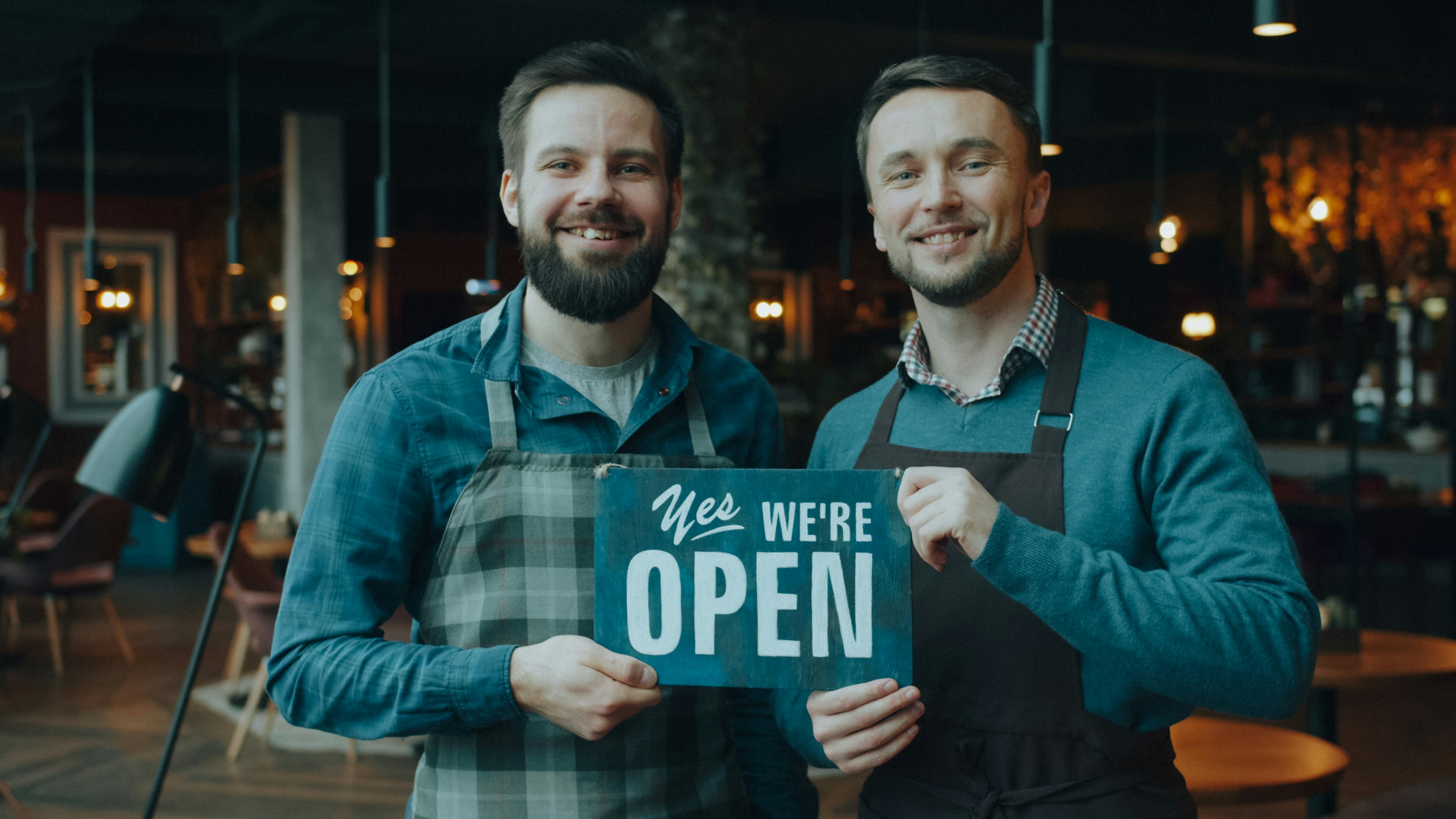 Two men in aprons holding a sign reading "YES WE'RE OPEN" in a restaurant