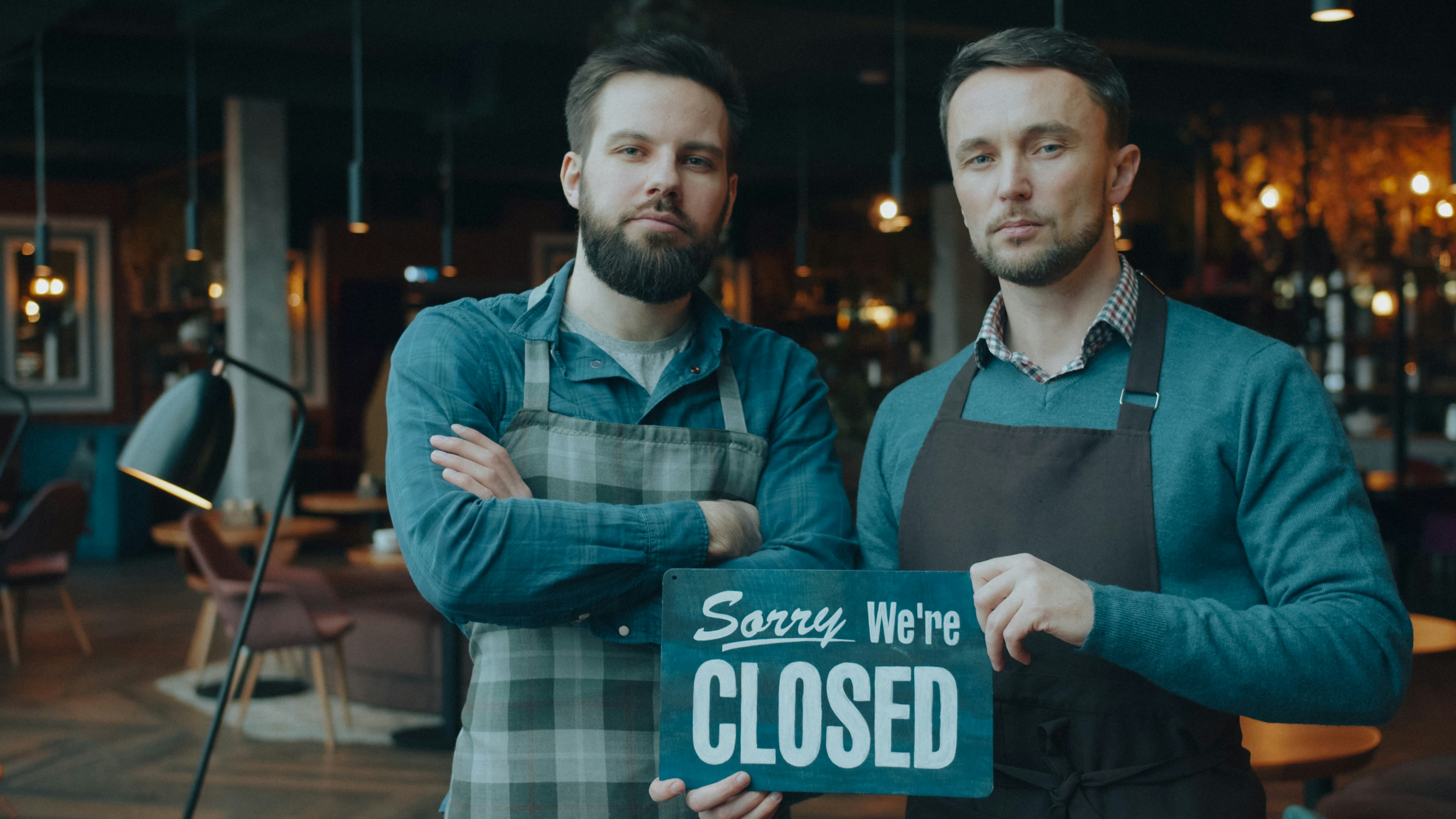 Two men in aprons holding a sign reading "Sorry We're CLOSED" in a restaurant