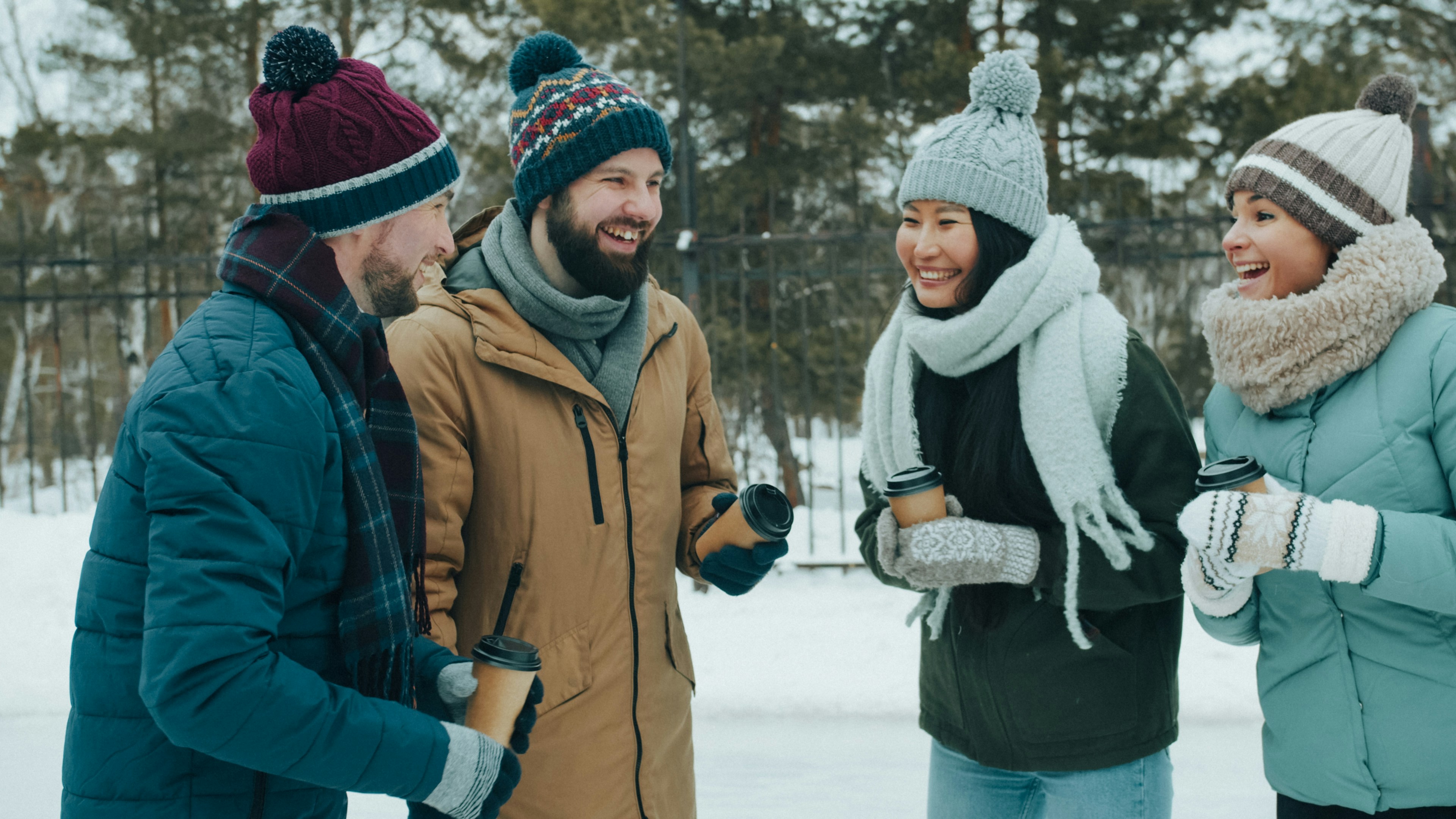 Four people in winter clothing holding coffee cups while laughing in a snowy park