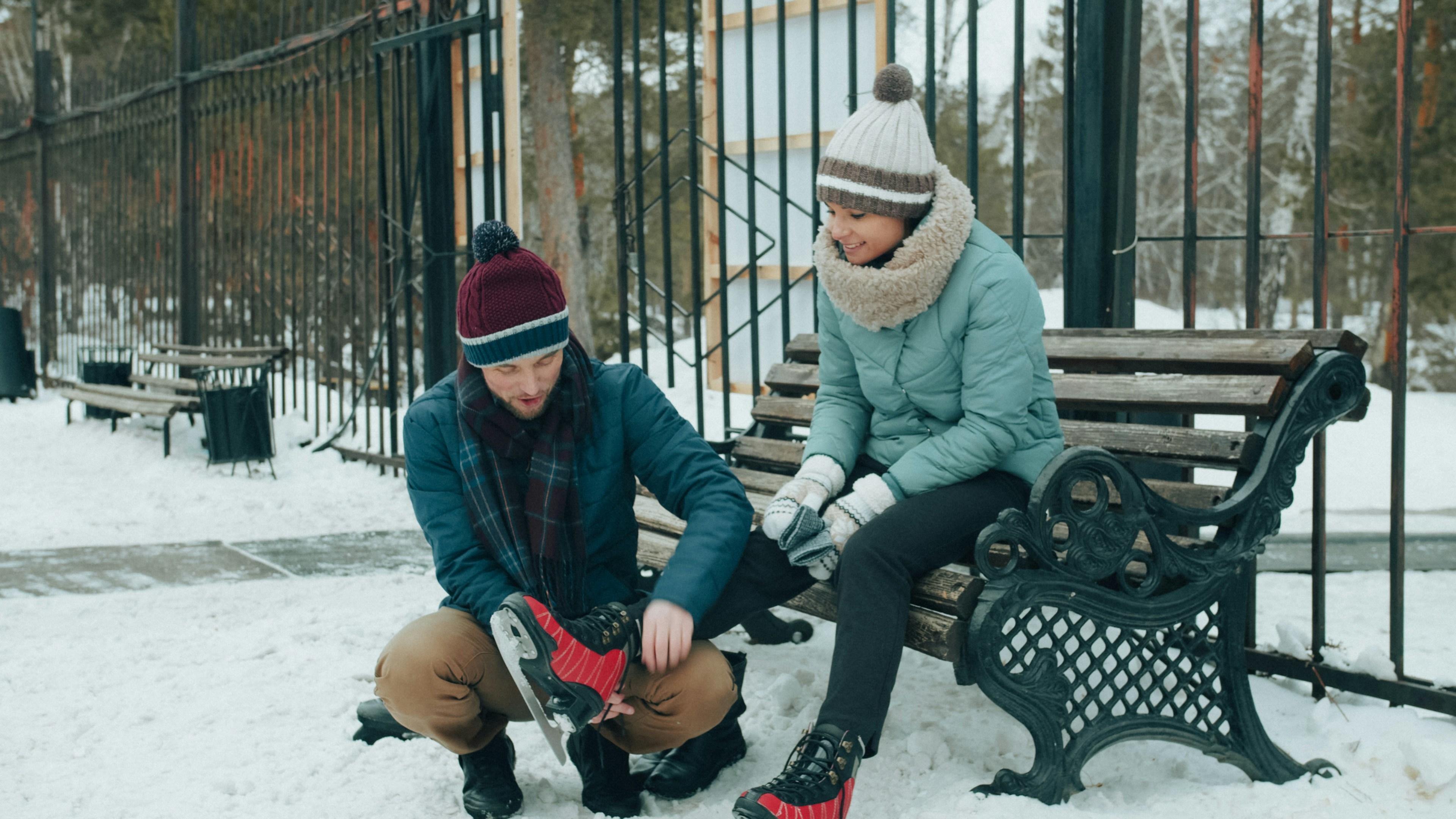 A man helping a woman put on ice skates on a snowy park bench