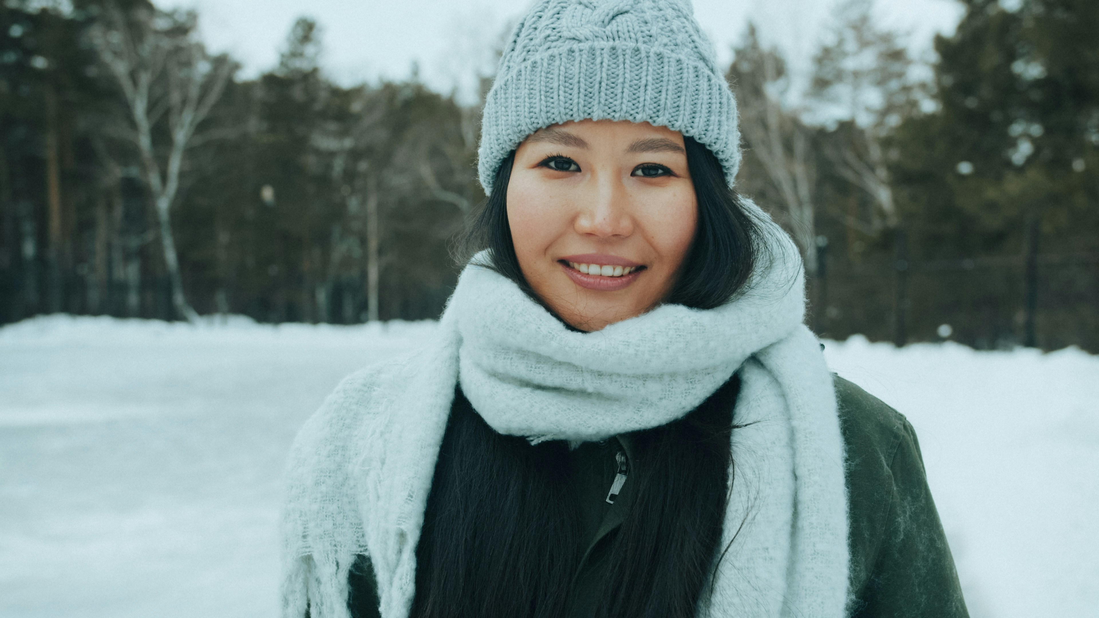 A woman wearing a grey knit hat and scarf in a snowy forest