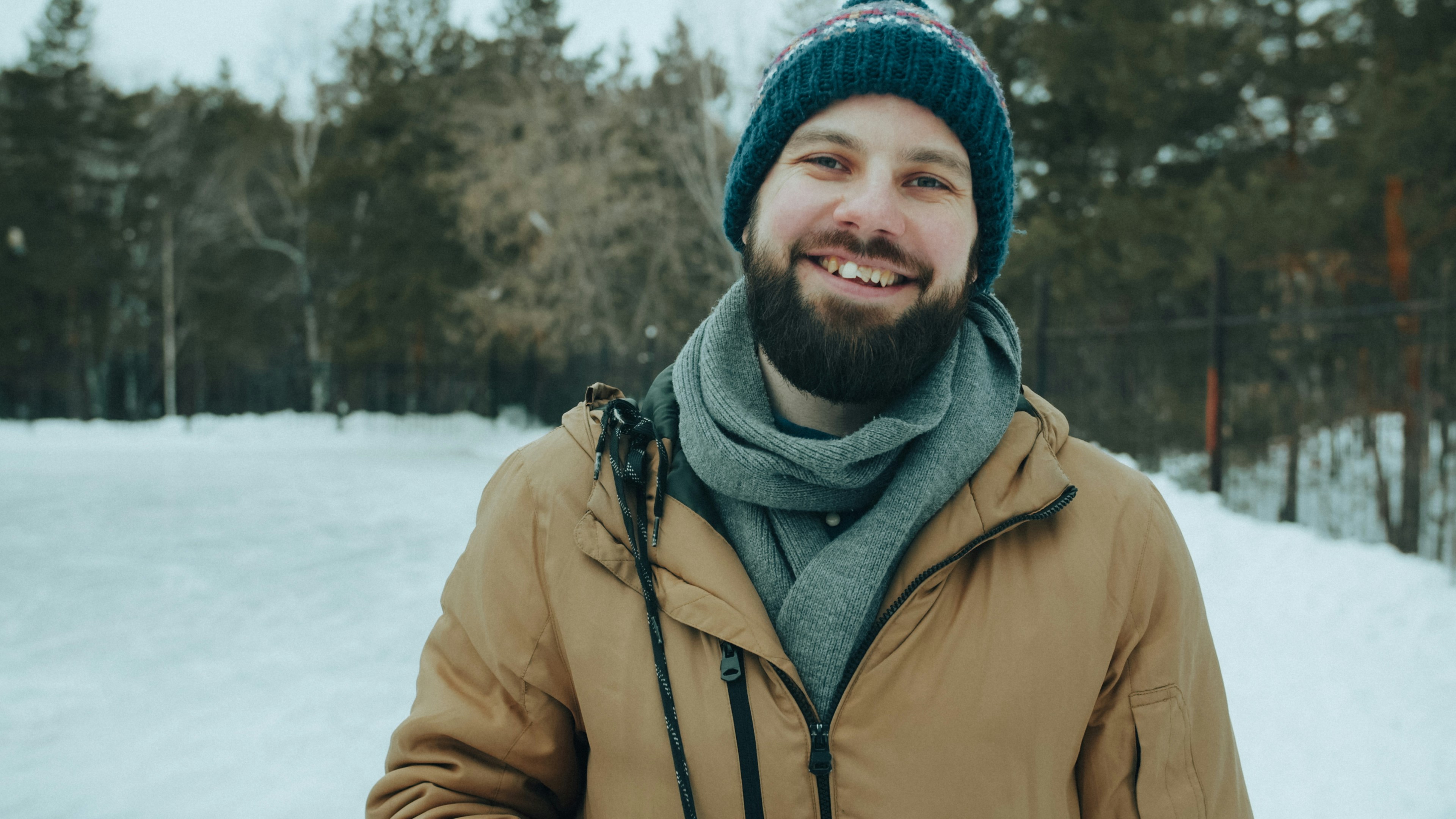 A smiling man with a beard wearing a tan jacket and blue knit hat