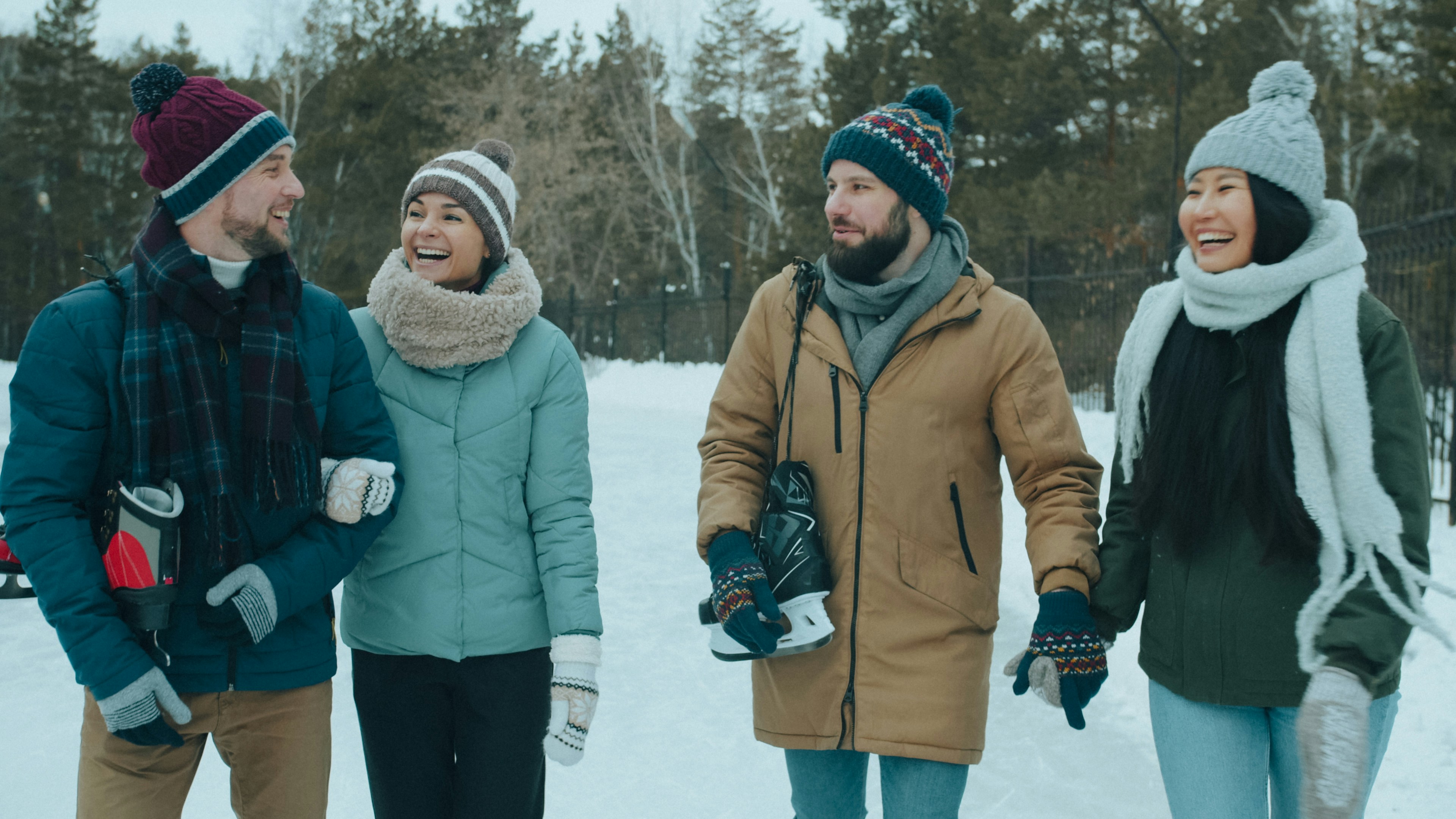 Four adults in winter clothing laughing while carrying ice skates on a snowy path