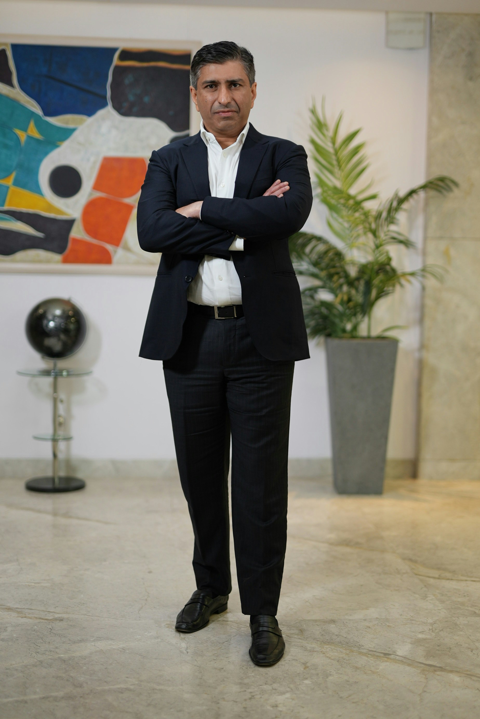 A man in a dark suit and white shirt with arms crossed in an office