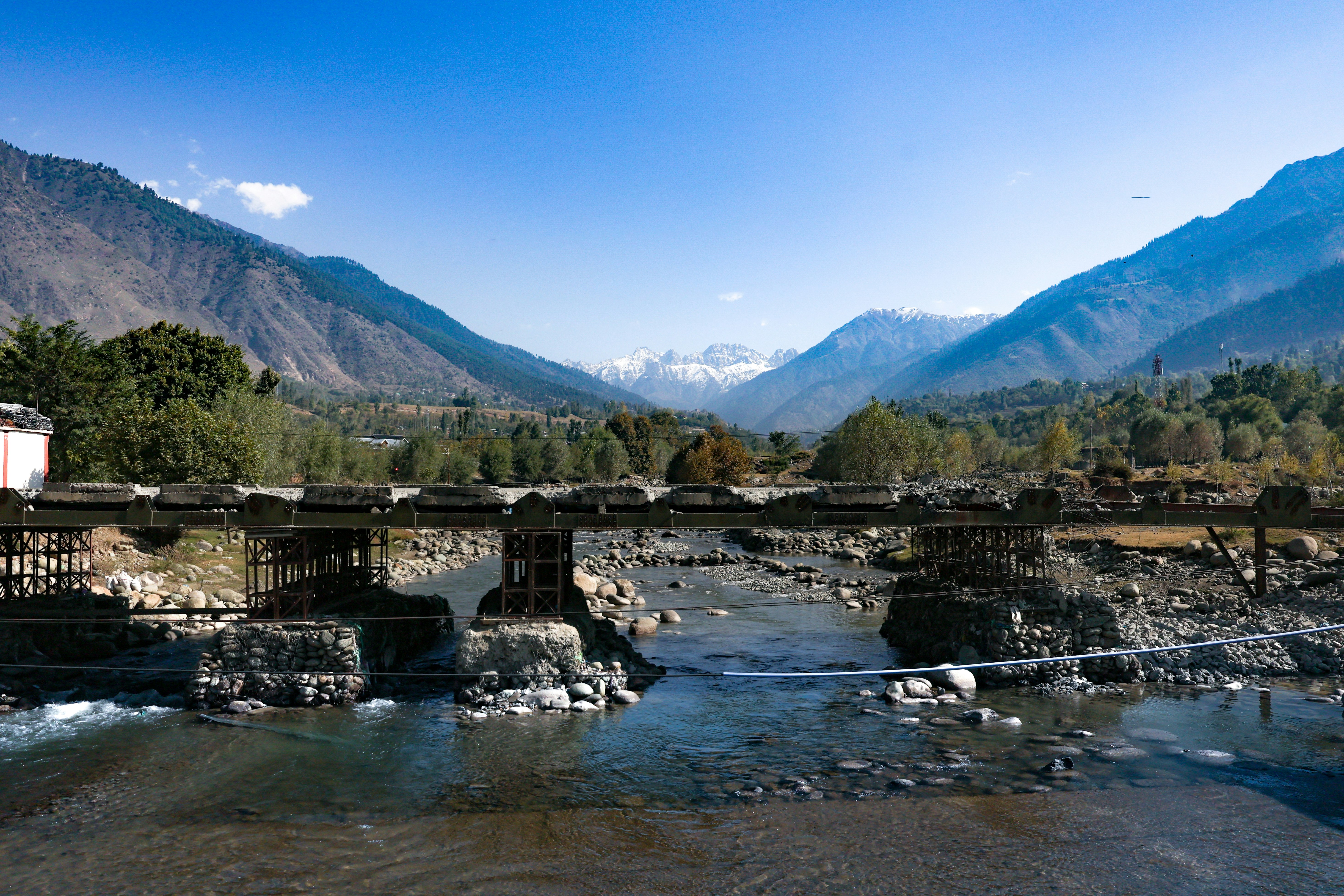 A metal bridge over a rocky river with snow-capped mountains in the distance