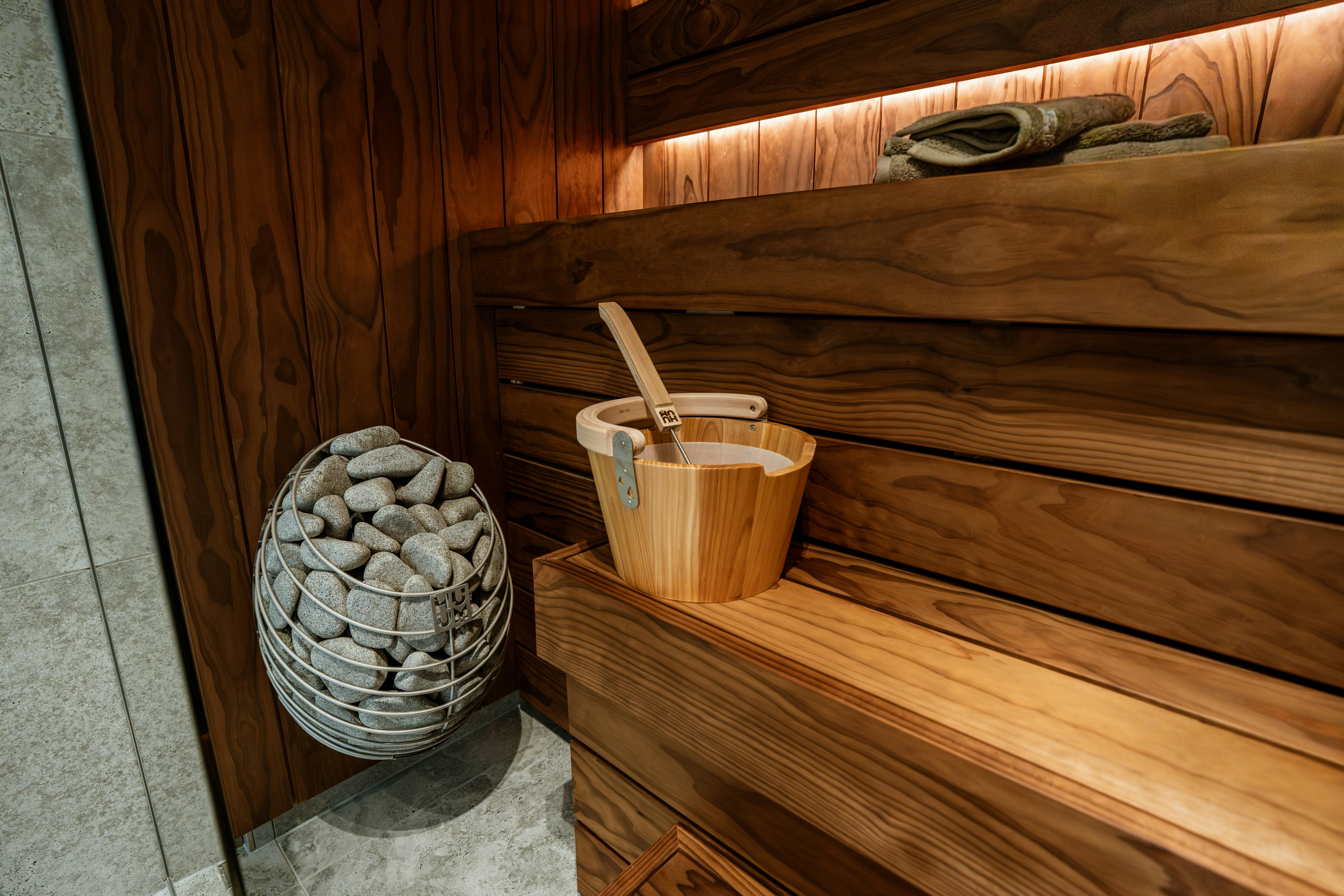 A wooden sauna bucket with a ladle on a bench beside a stone heater