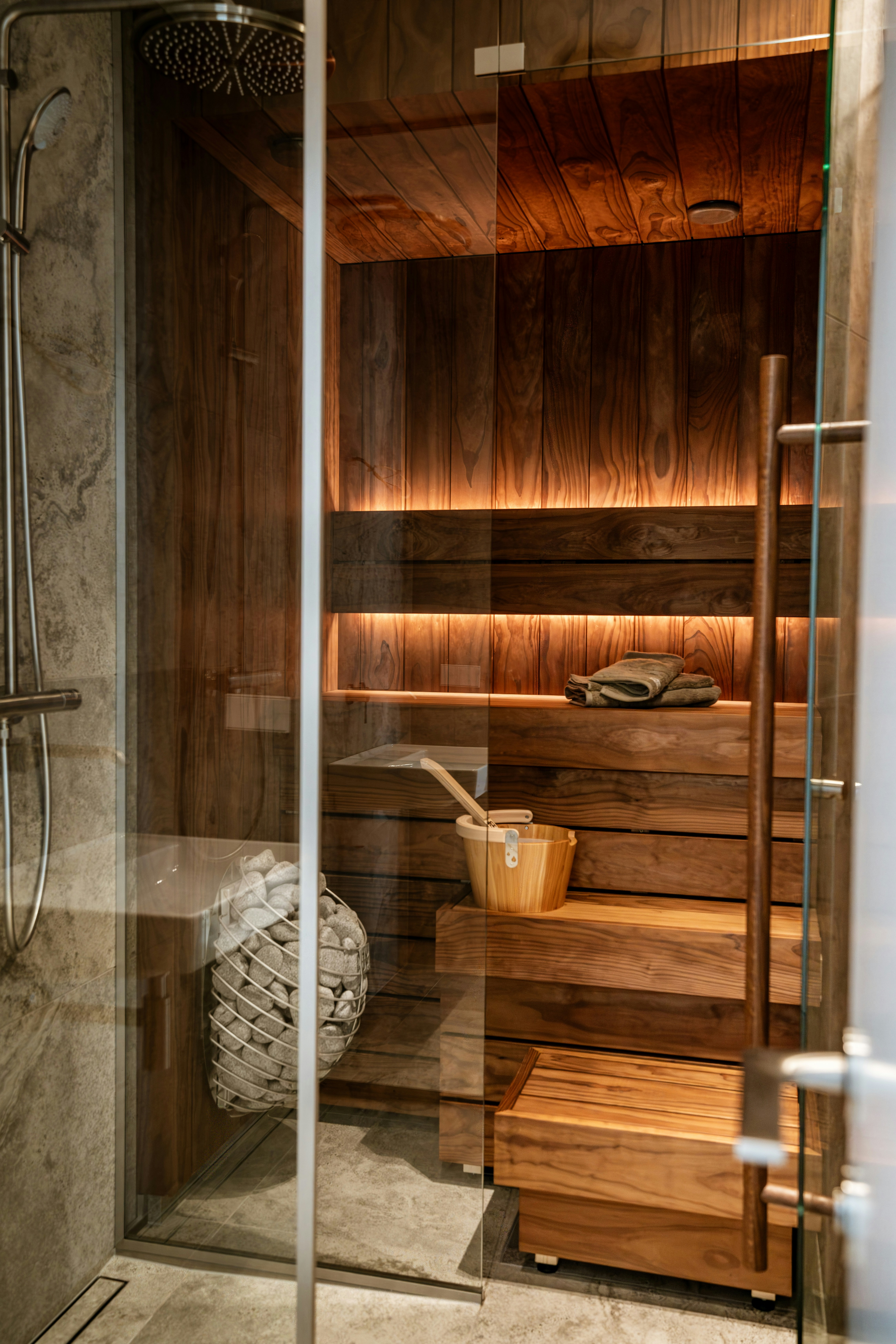 A wooden sauna with tiered benches, a bucket, and stones behind glass