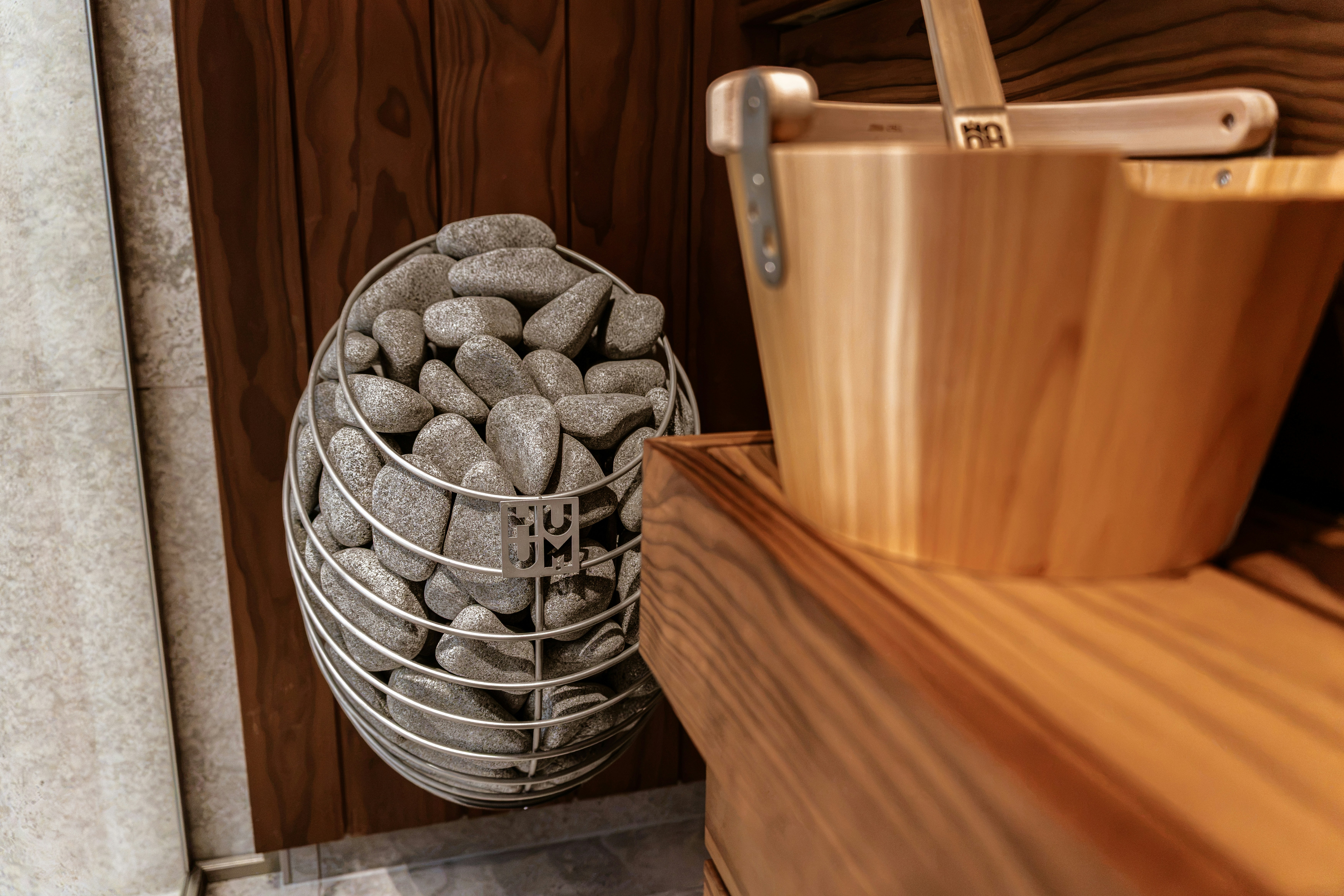A metal cage filled with grey sauna stones beside a wooden bucket