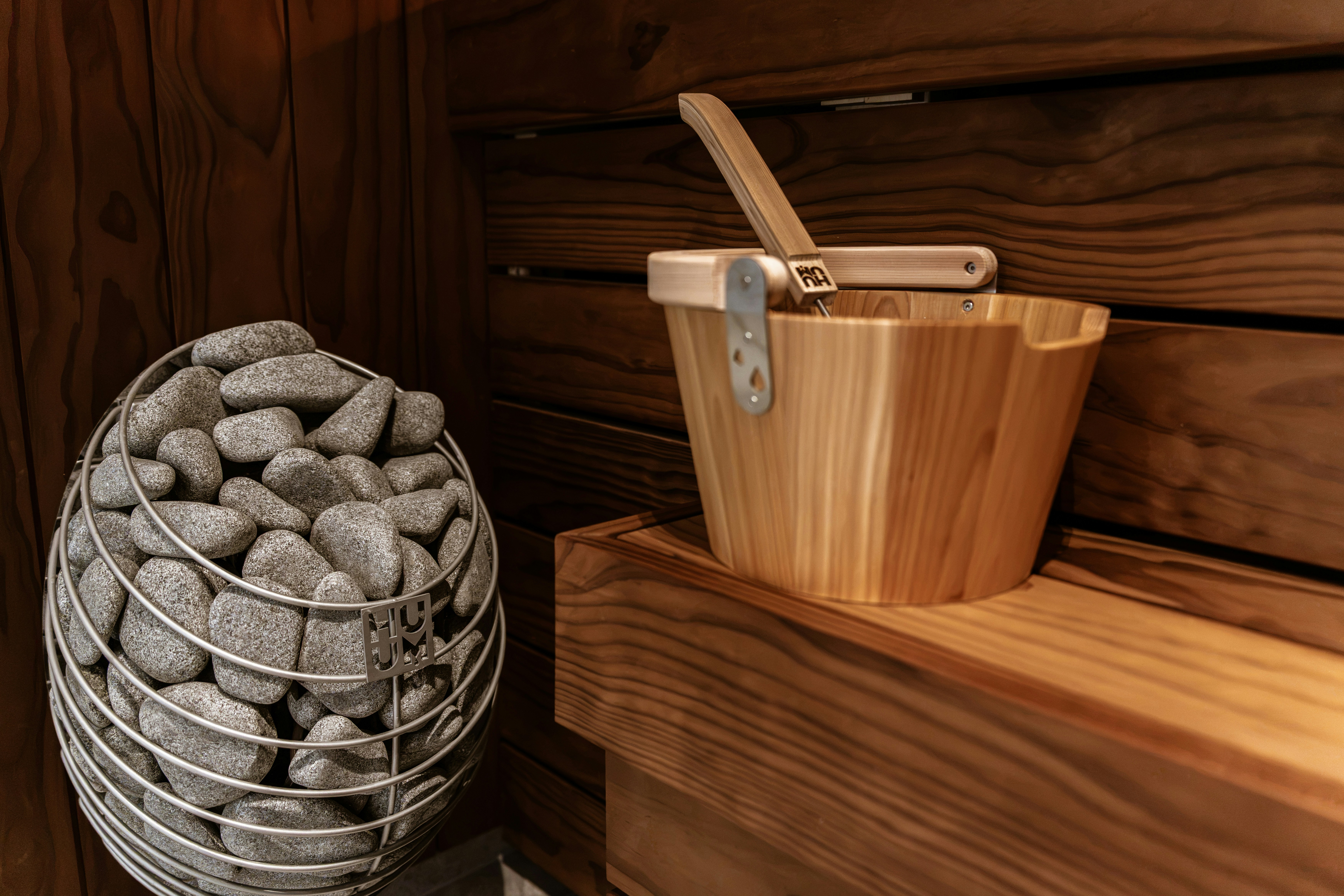 A wooden sauna bucket with a ladle beside a wire basket of stones