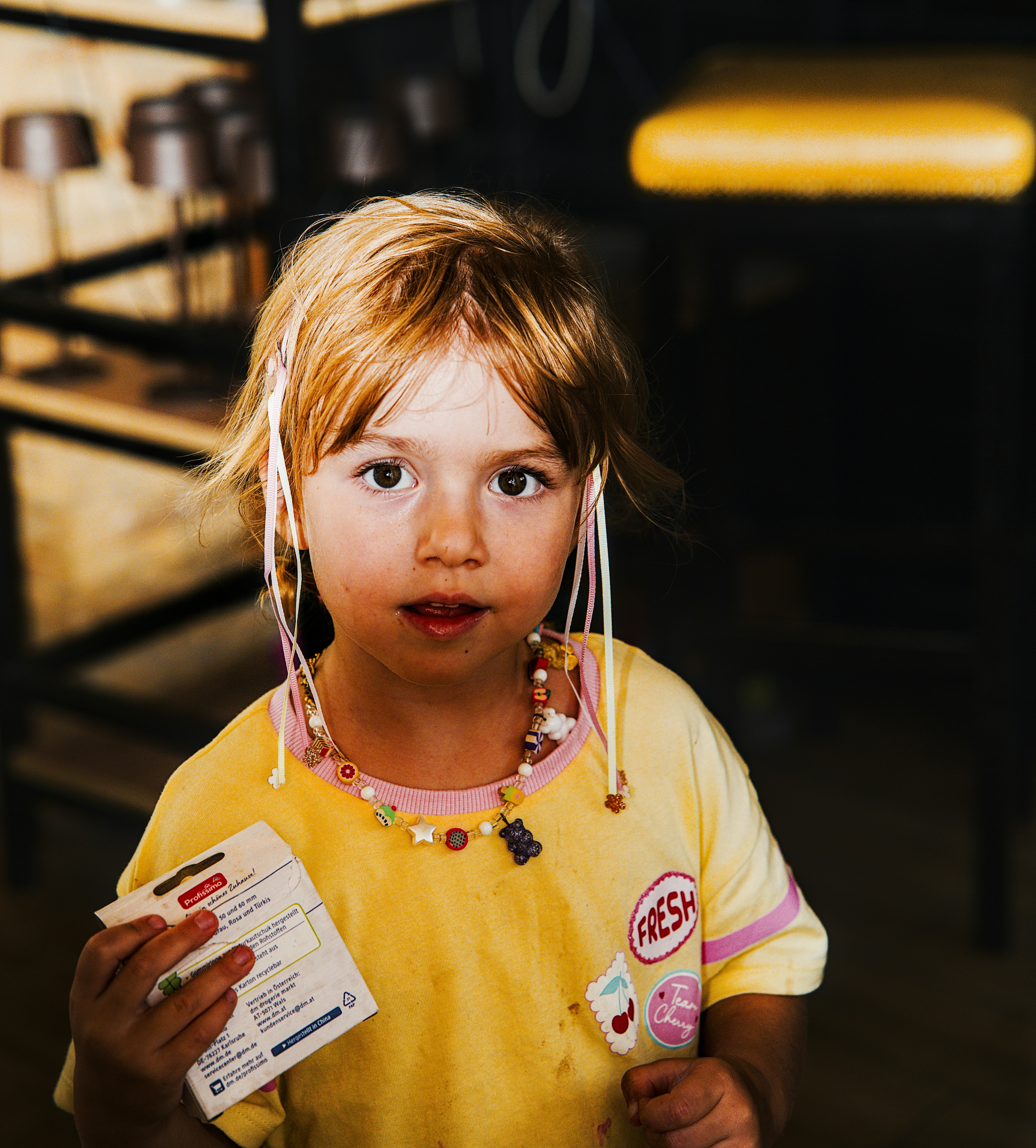 inquisitive: A child with blonde hair and a beaded necklace holding a small product box