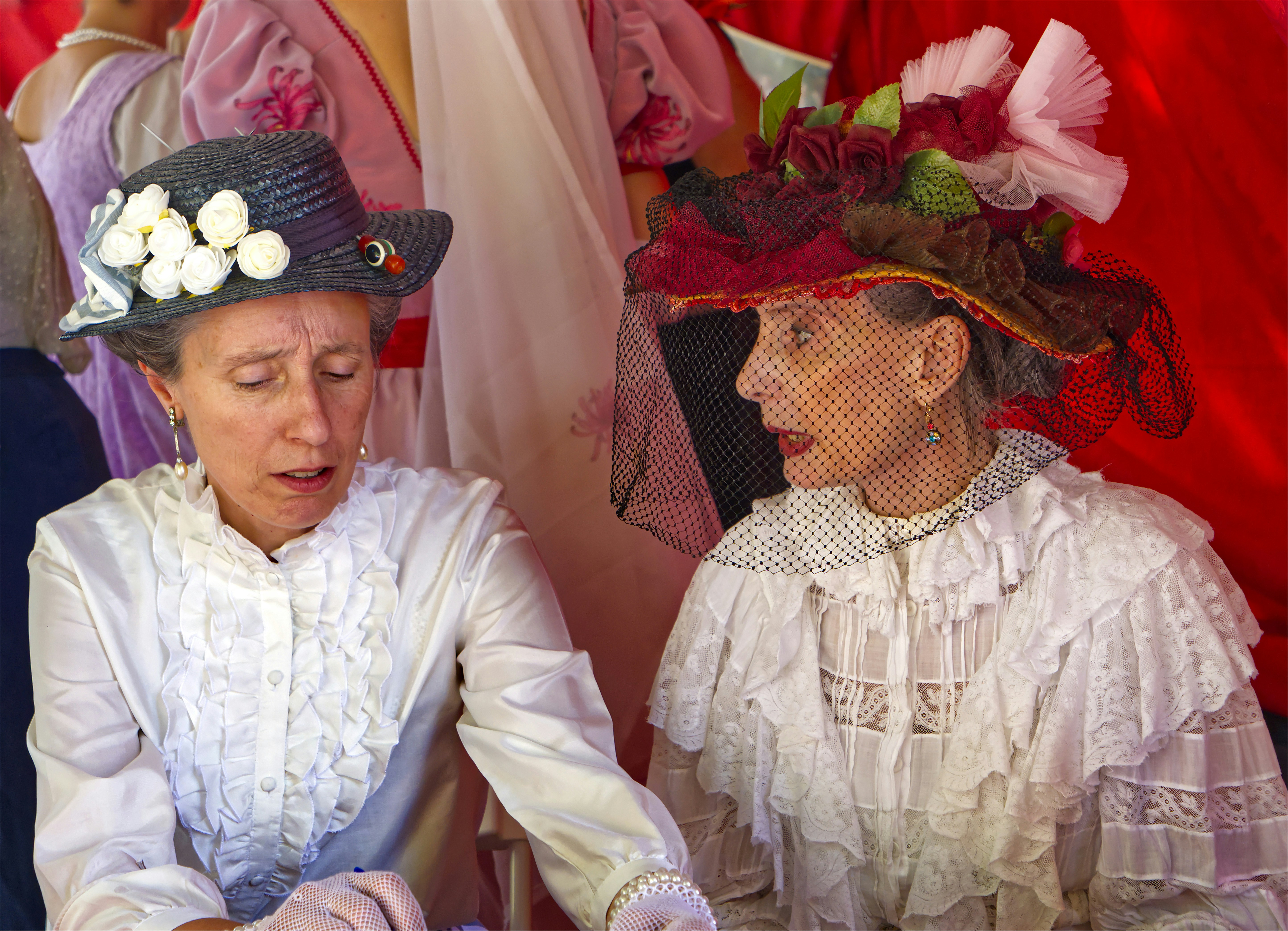 Two women in vintage white ruffled blouses and ornate hats talking together