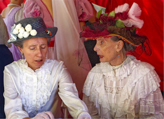 Two women in vintage white ruffled blouses and ornate hats talking together