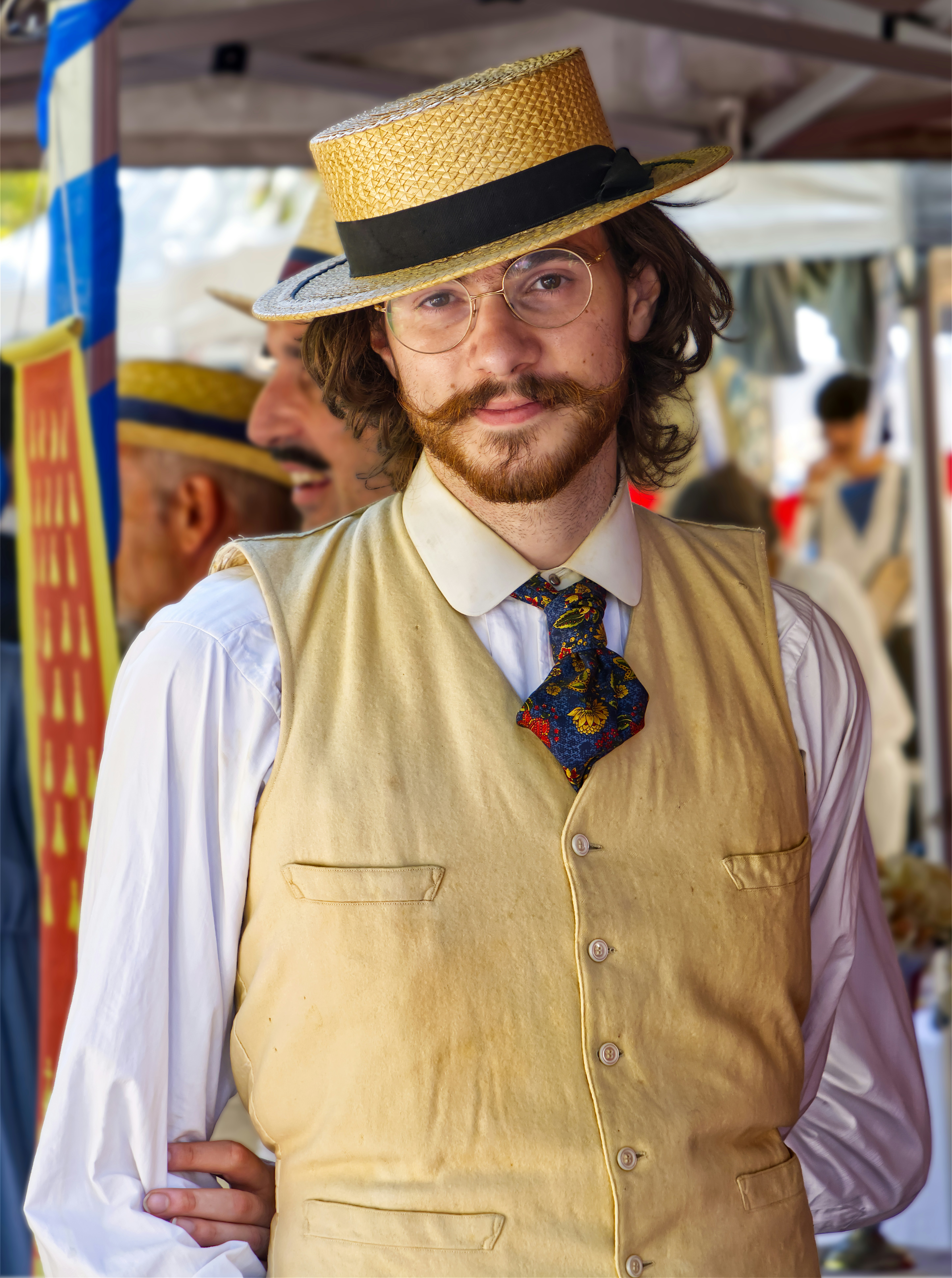 A man with a mustache wearing a straw boater hat, glasses, and a tan vest