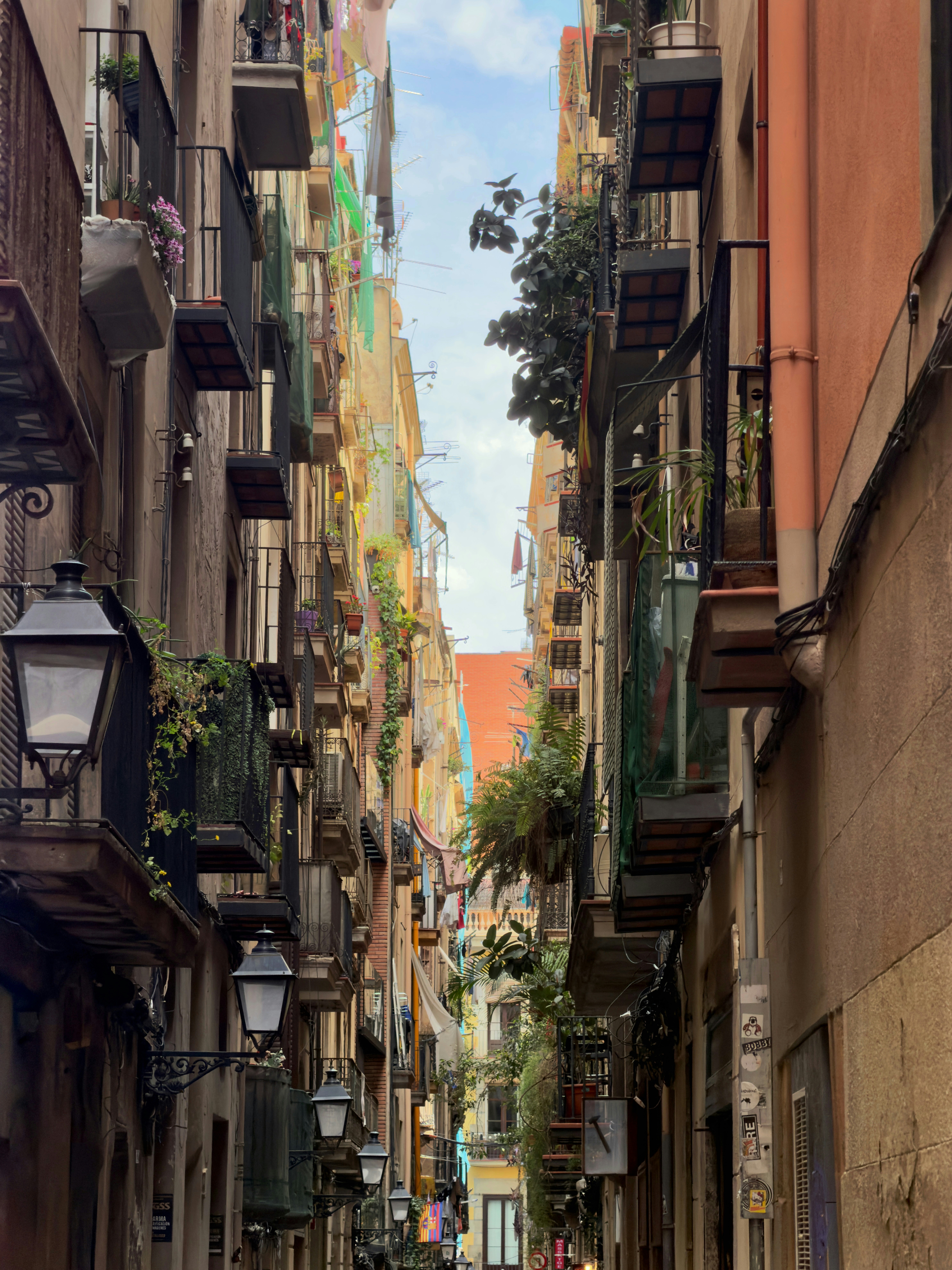 A narrow street between tall apartment buildings with balconies and hanging plants in Barcelona