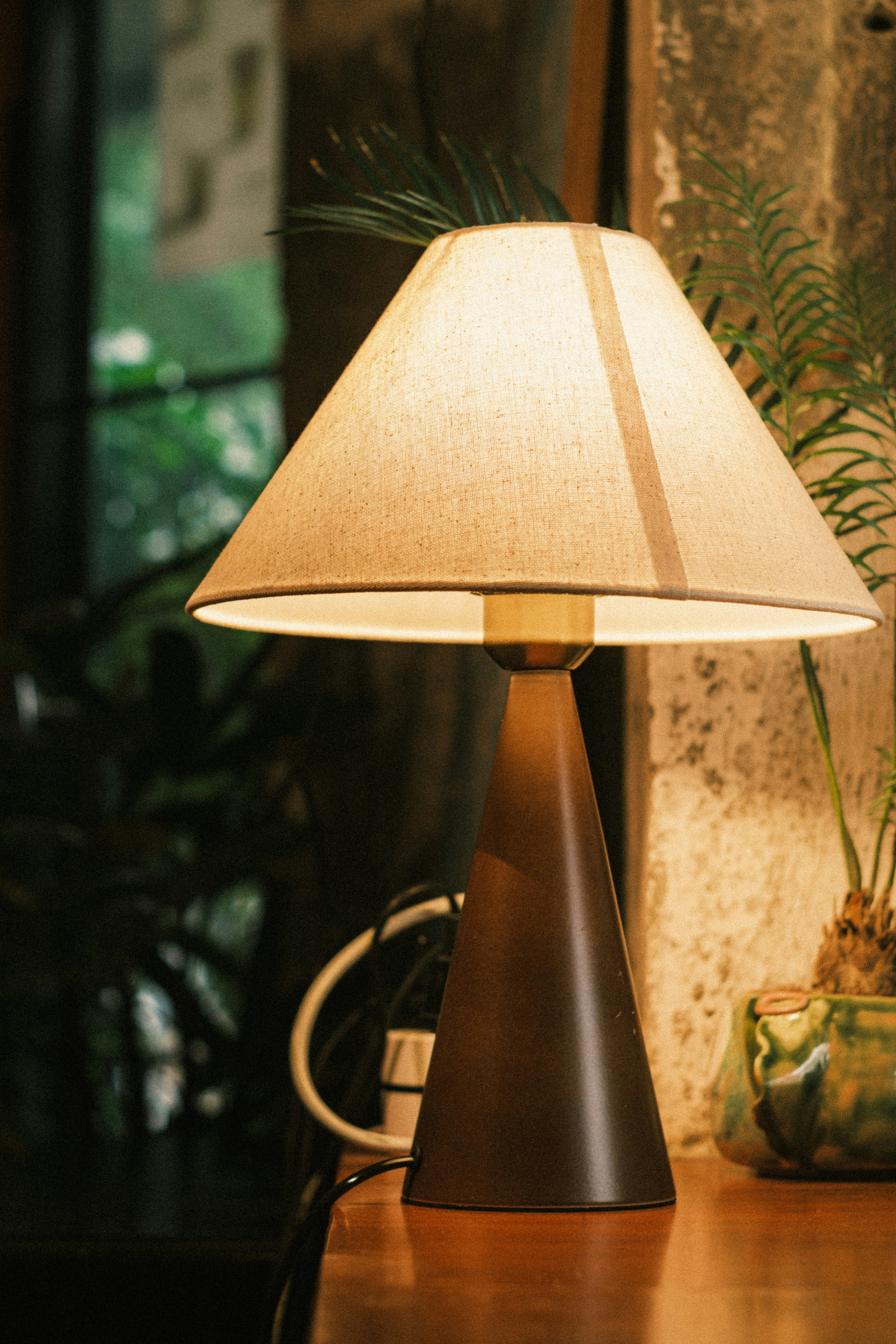 A lit table lamp with a conical base and fabric shade on a wooden surface
