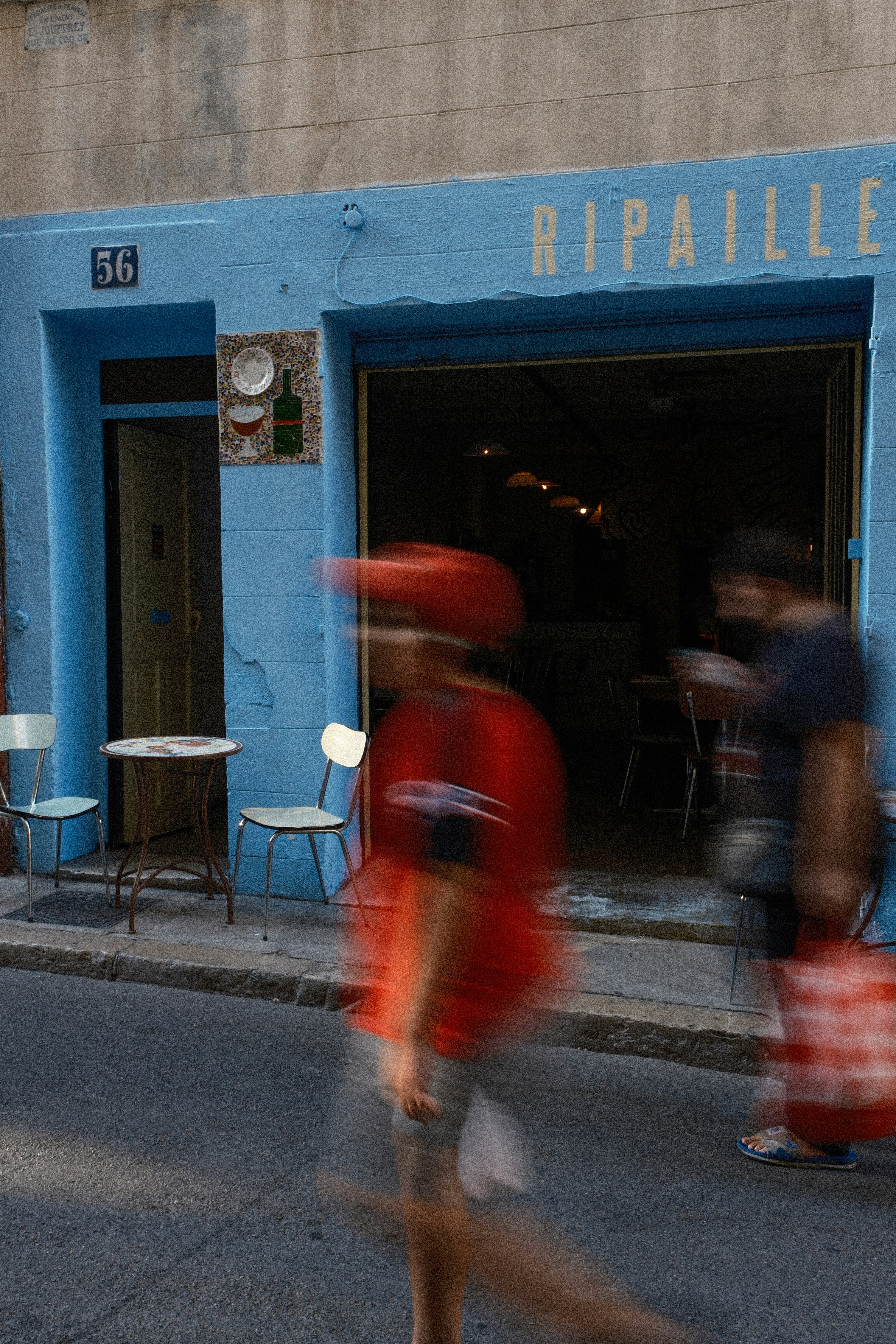 Two blurred people walking past a blue restaurant facade with outdoor seating