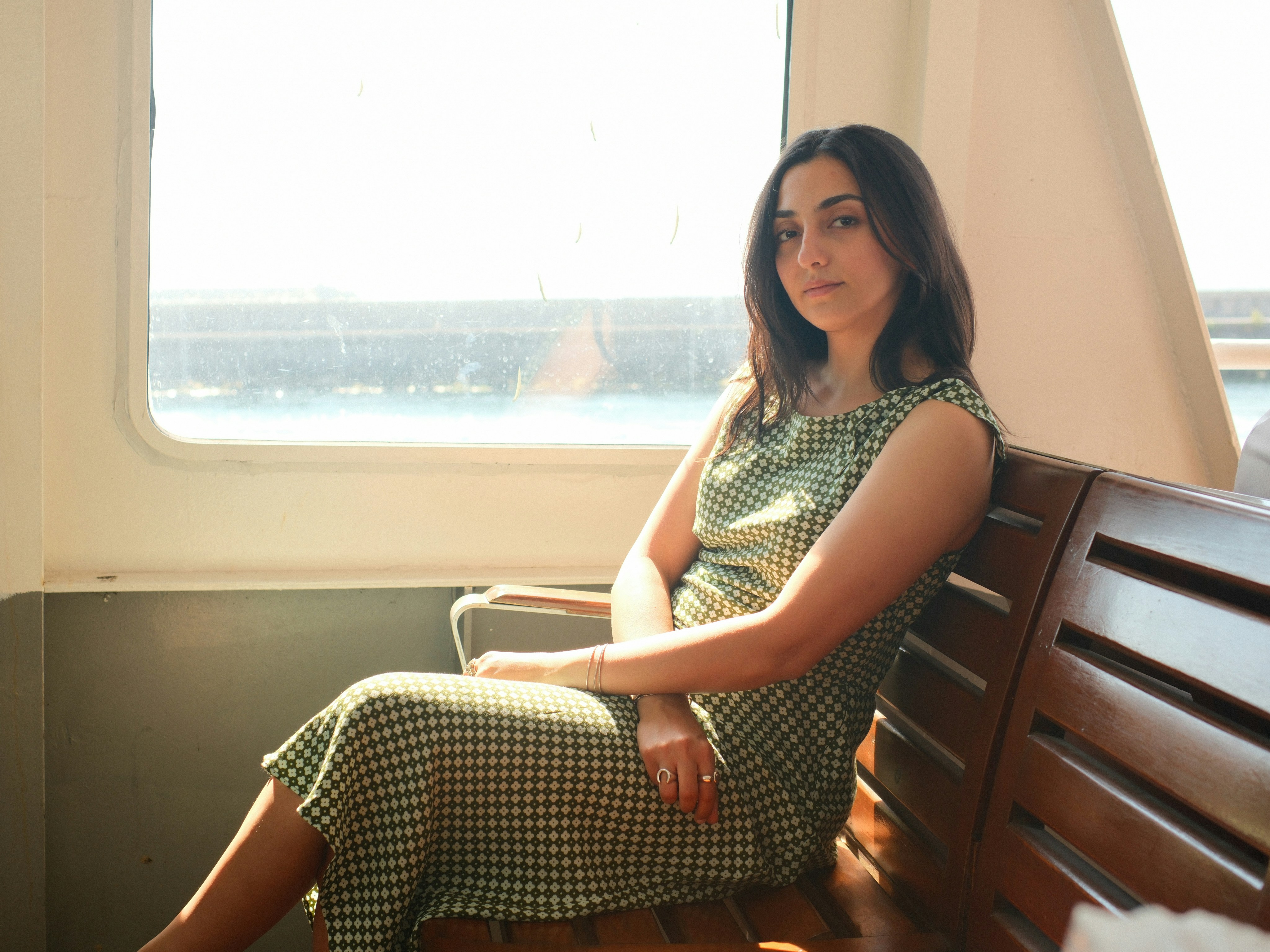 A woman in a green patterned dress sitting on a wooden bench by a window