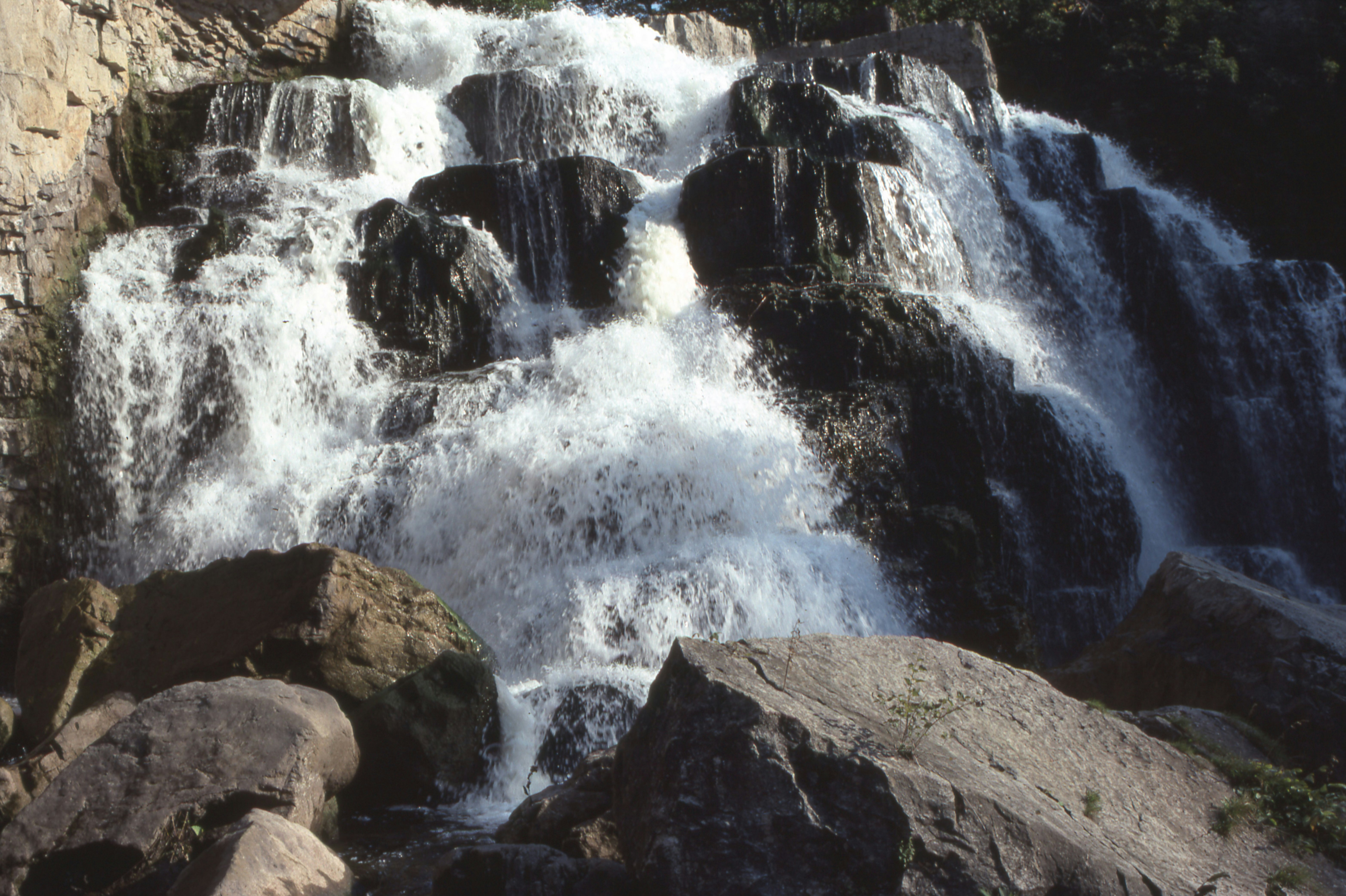 Water cascading over dark tiered rocks into a pool with large boulders