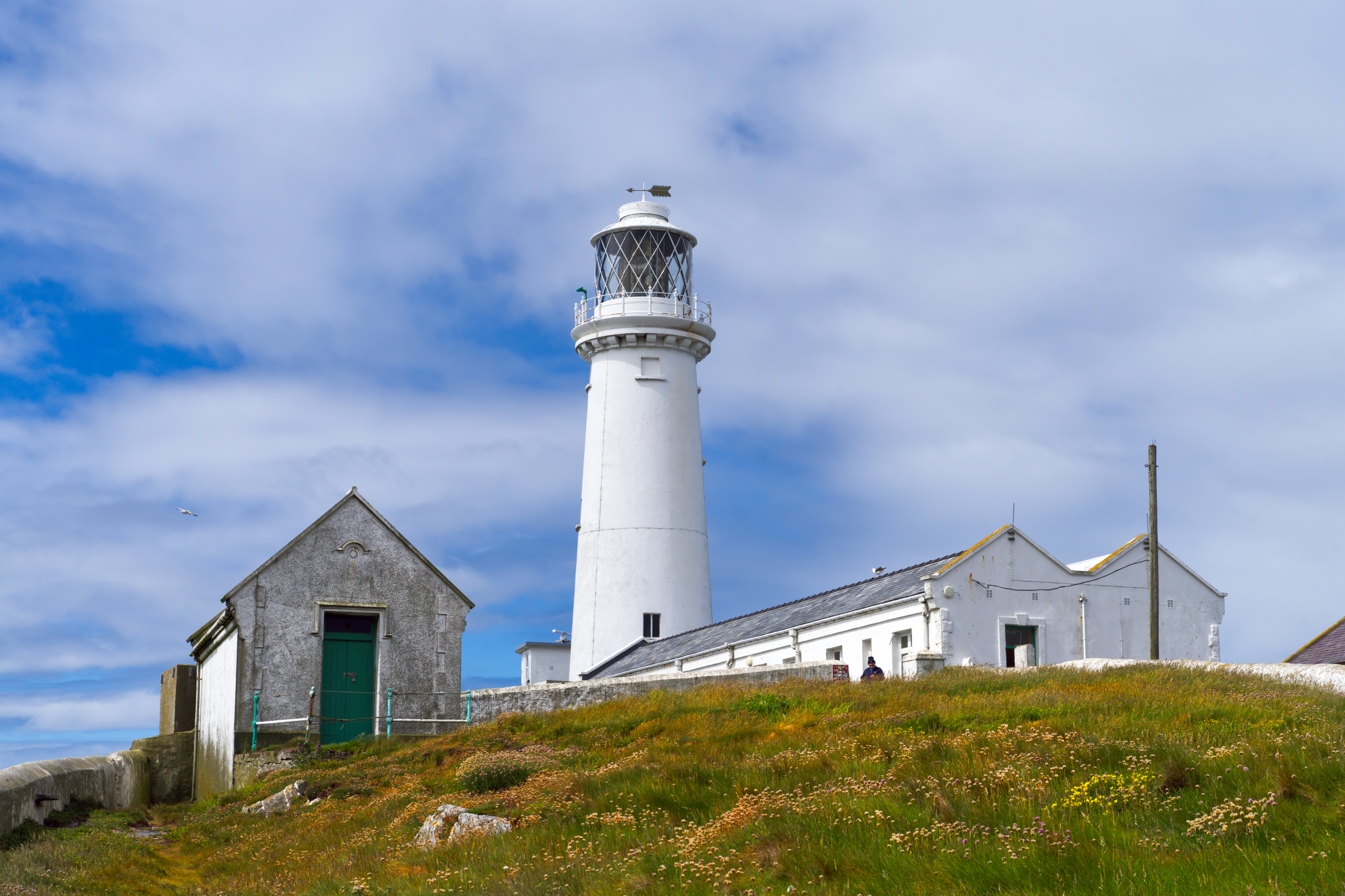 A white lighthouse and adjacent buildings on a grassy hill under a blue sky