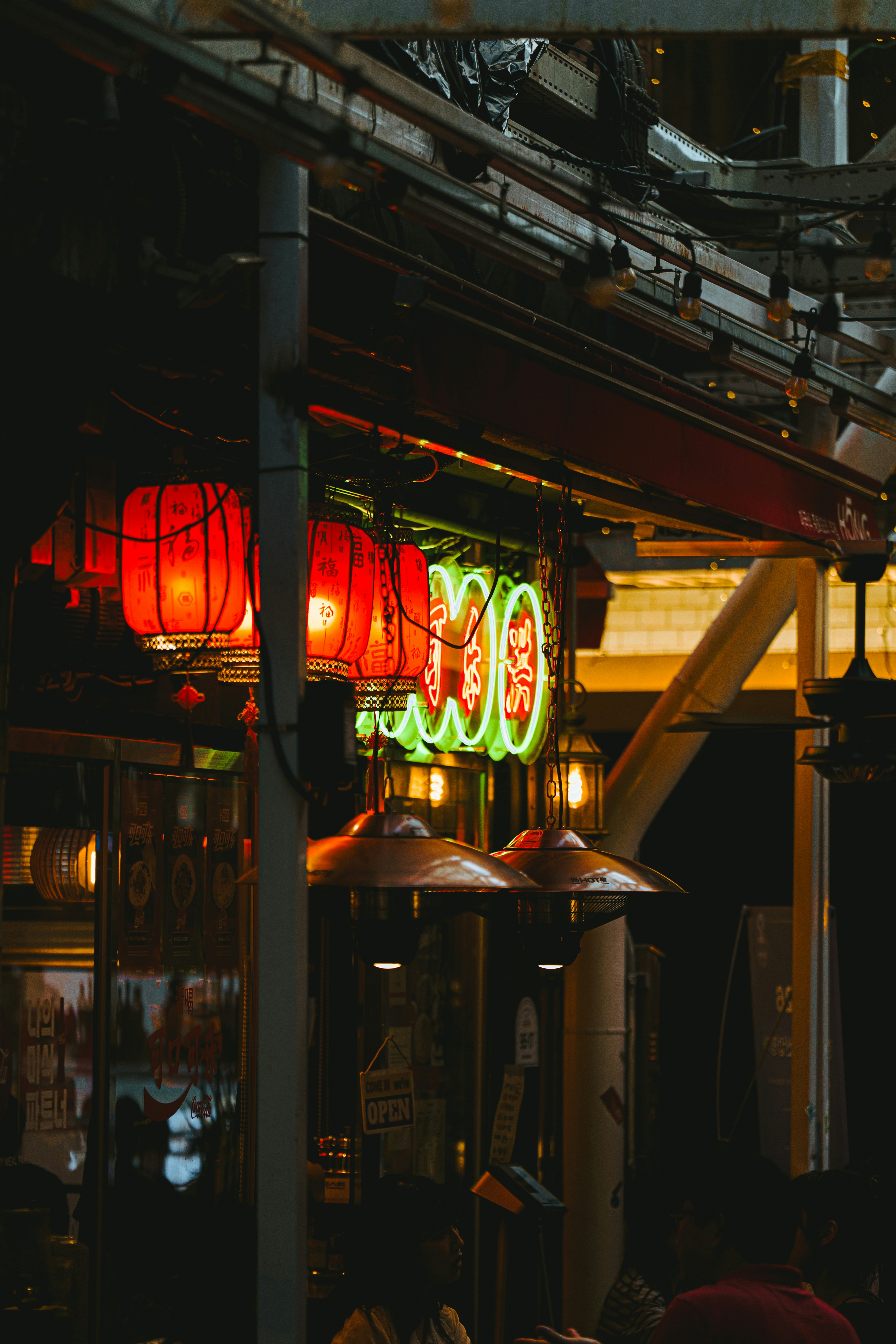 Red lanterns and neon signs glowing above outdoor heaters at a restaurant entrance