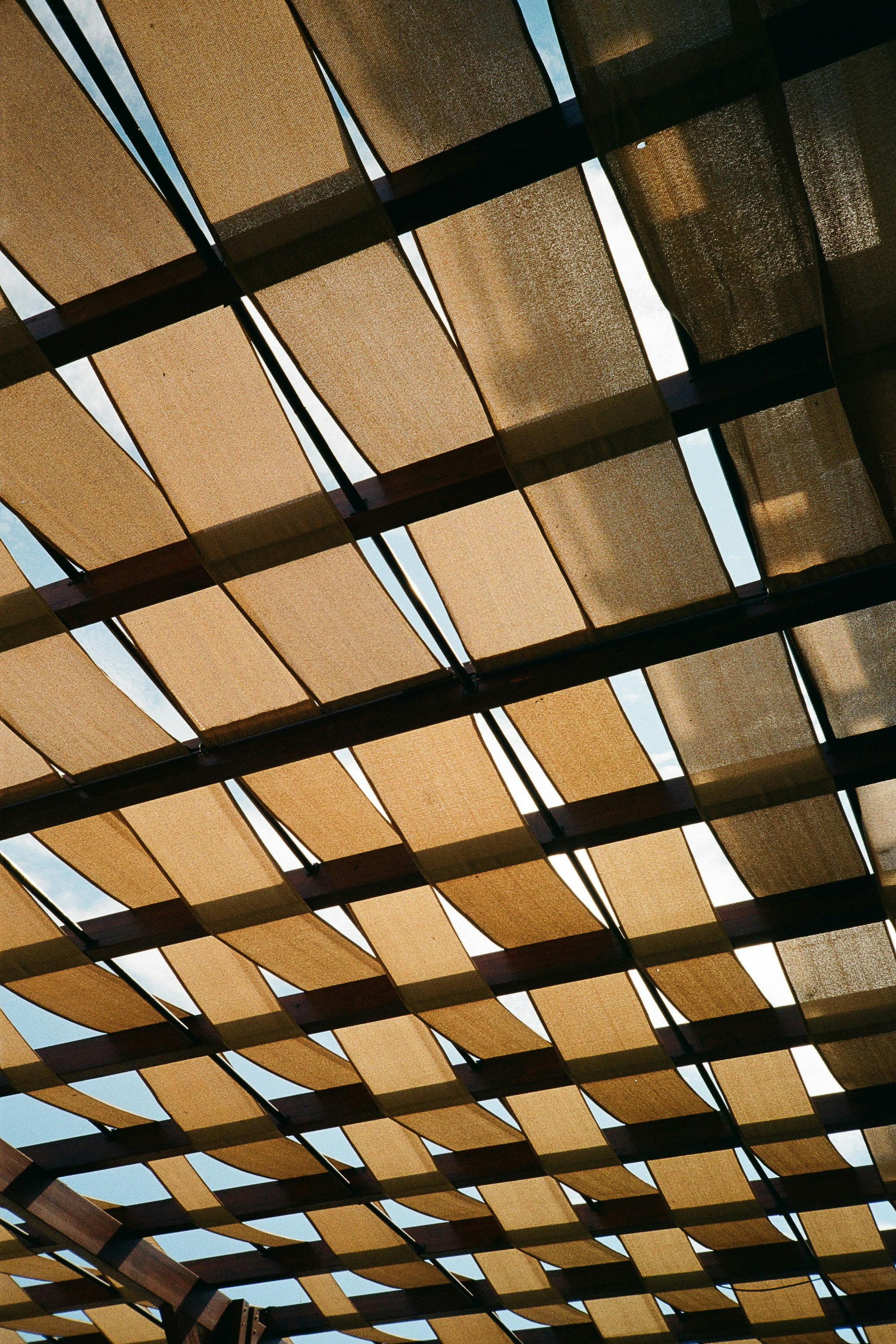 Tan fabric strips woven through a dark wooden pergola against a bright sky