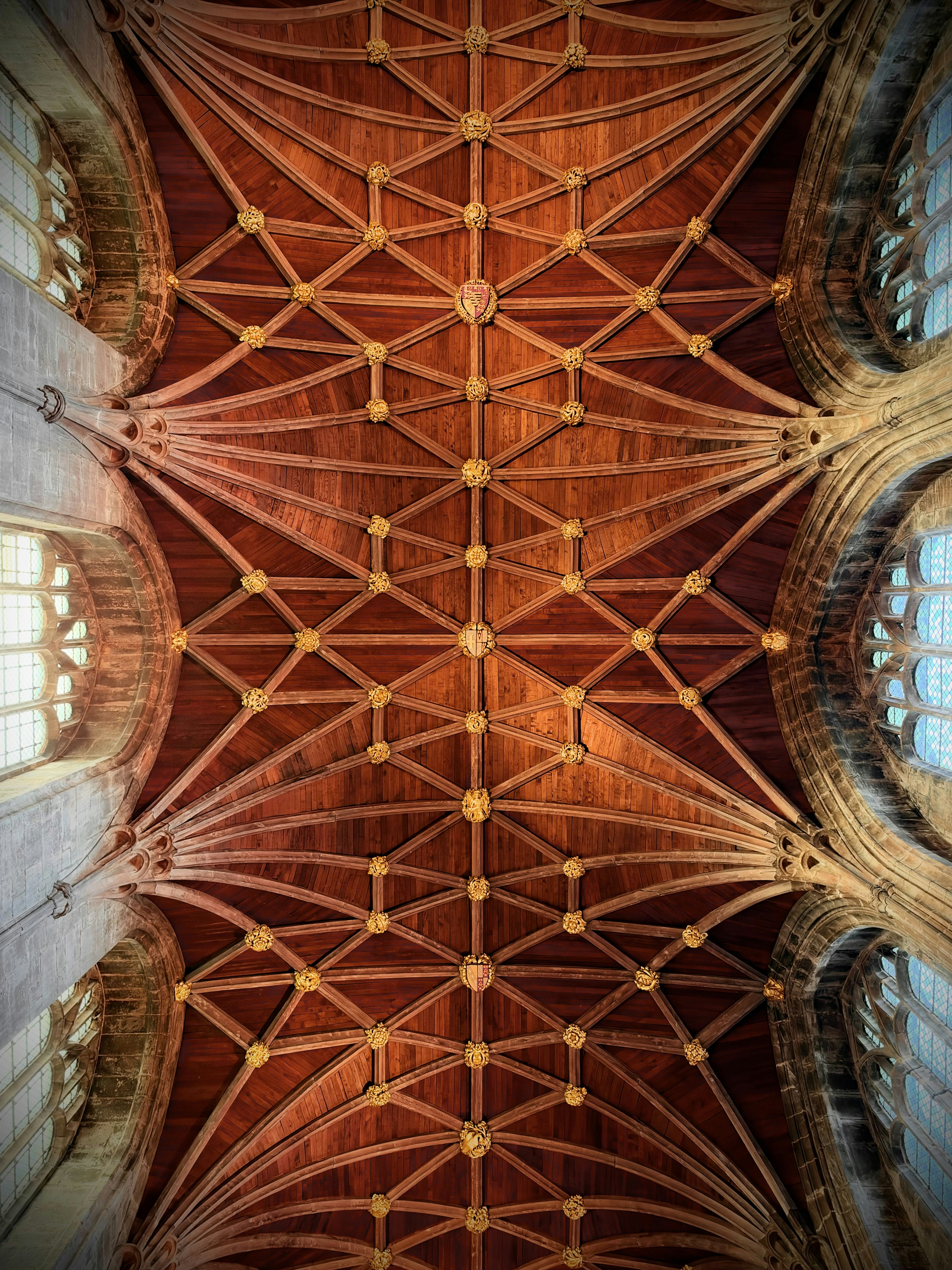 A wooden vaulted ceiling with intricate gold bosses and stone arched windows