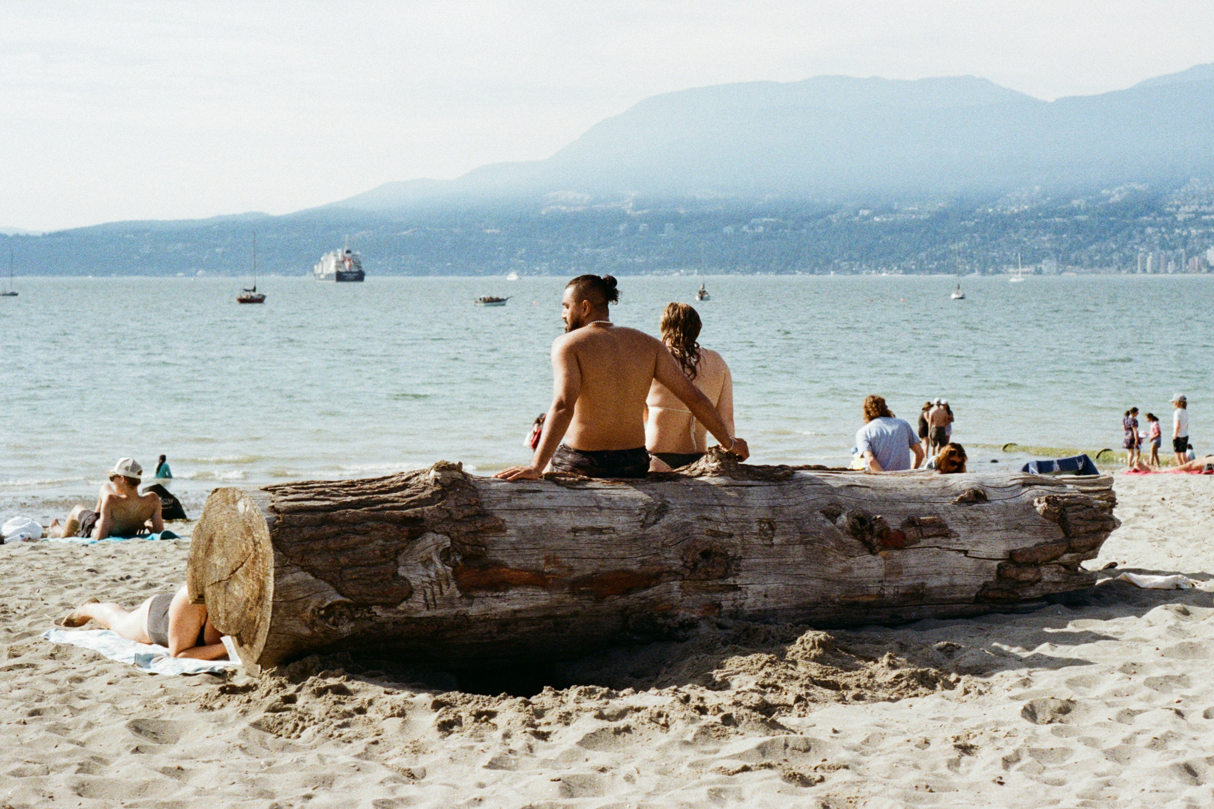 People relaxing on a large driftwood log on a sandy beach by the ocean