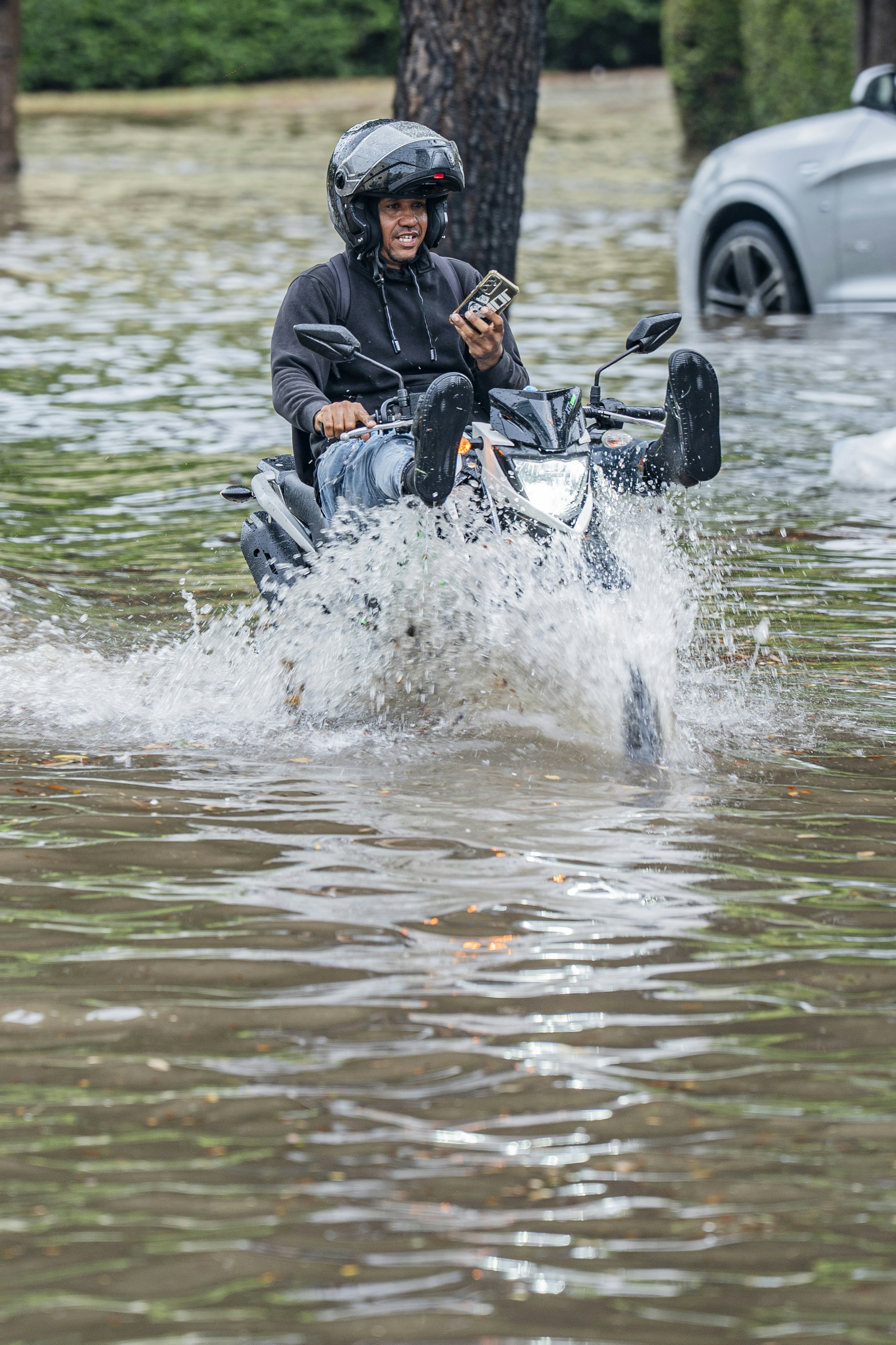 A person in a helmet riding a motorcycle through deep floodwater on a street