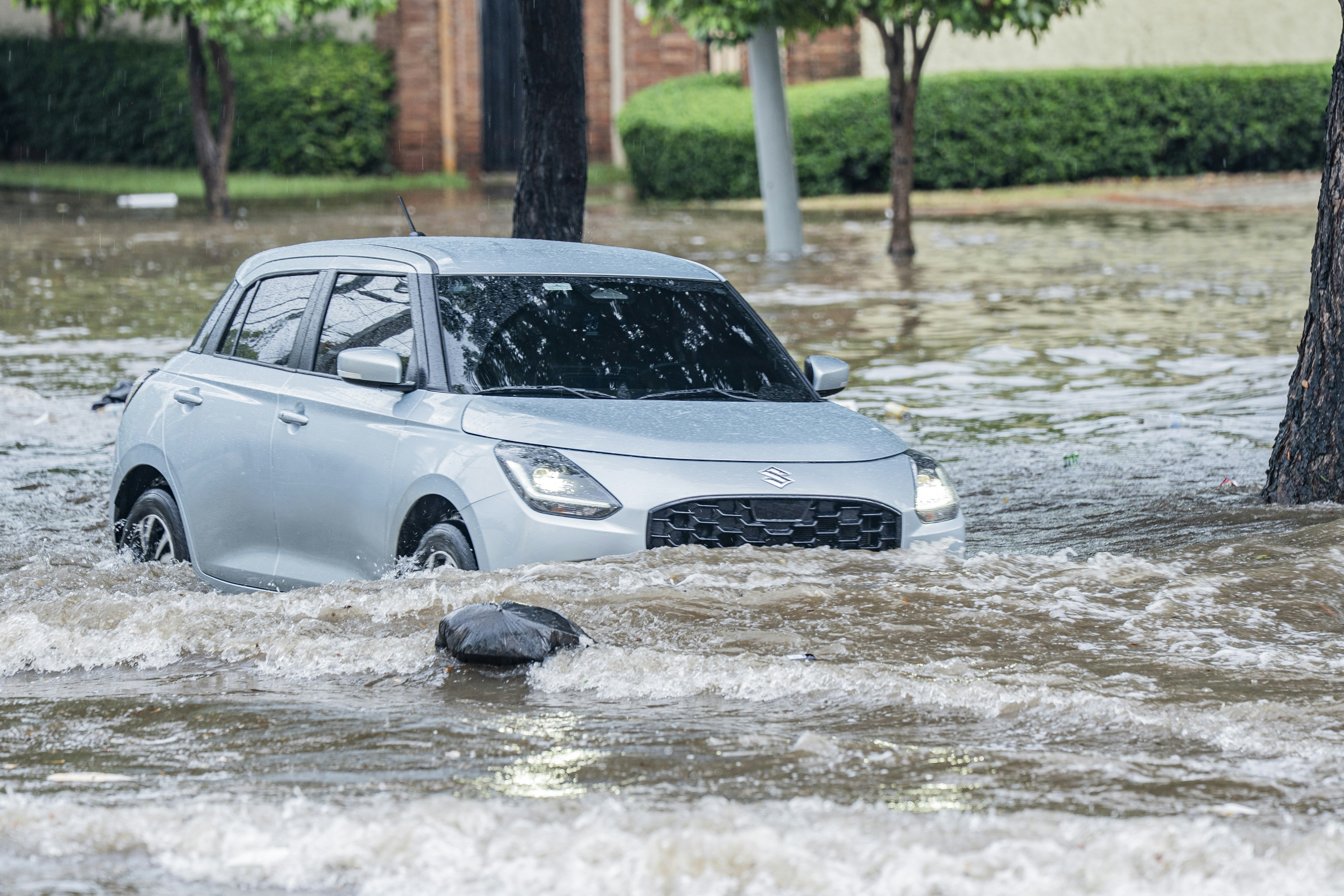 A silver hatchback driving through deep floodwater on a tree-lined street