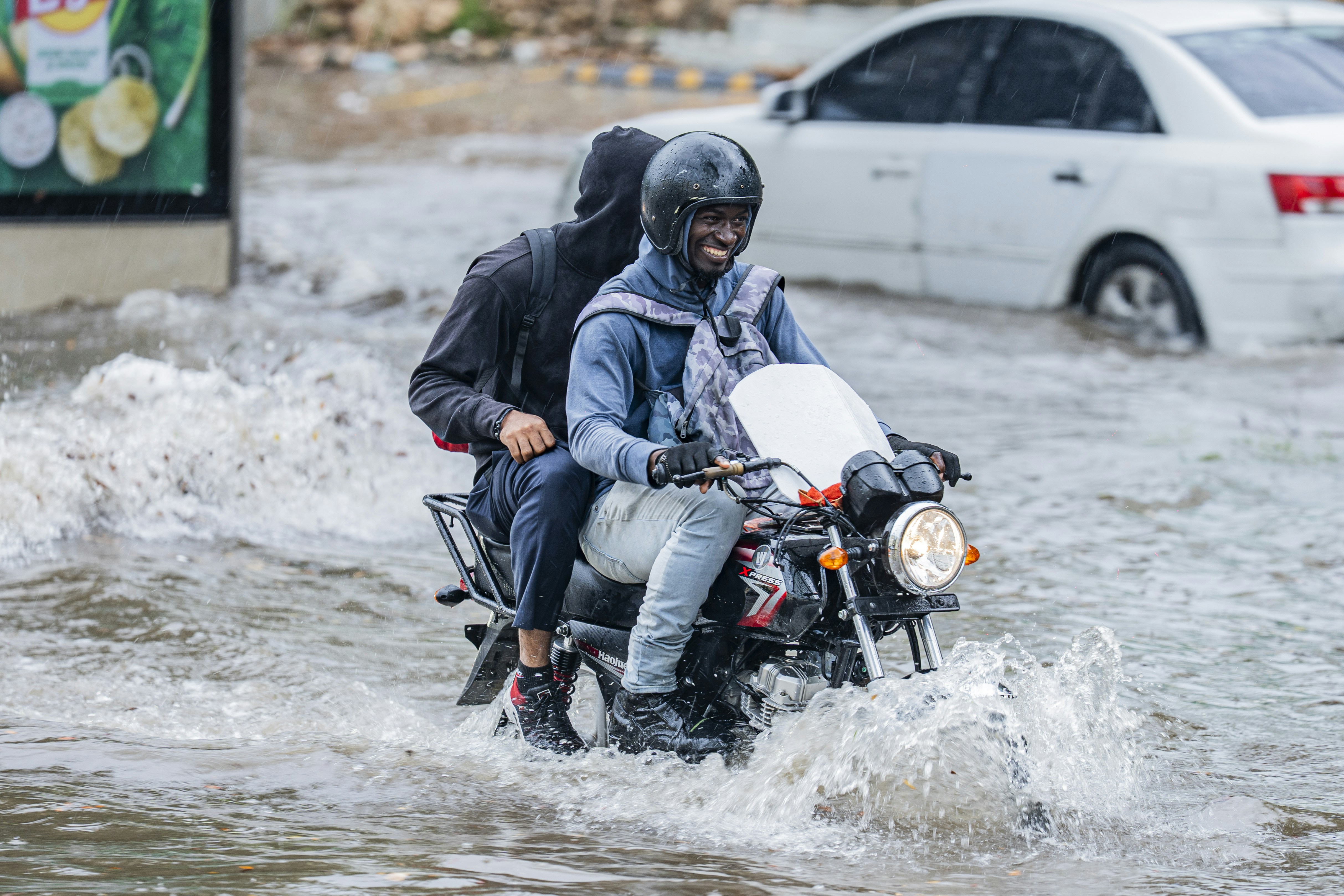 Two people riding a motorcycle through deep floodwater on a city street