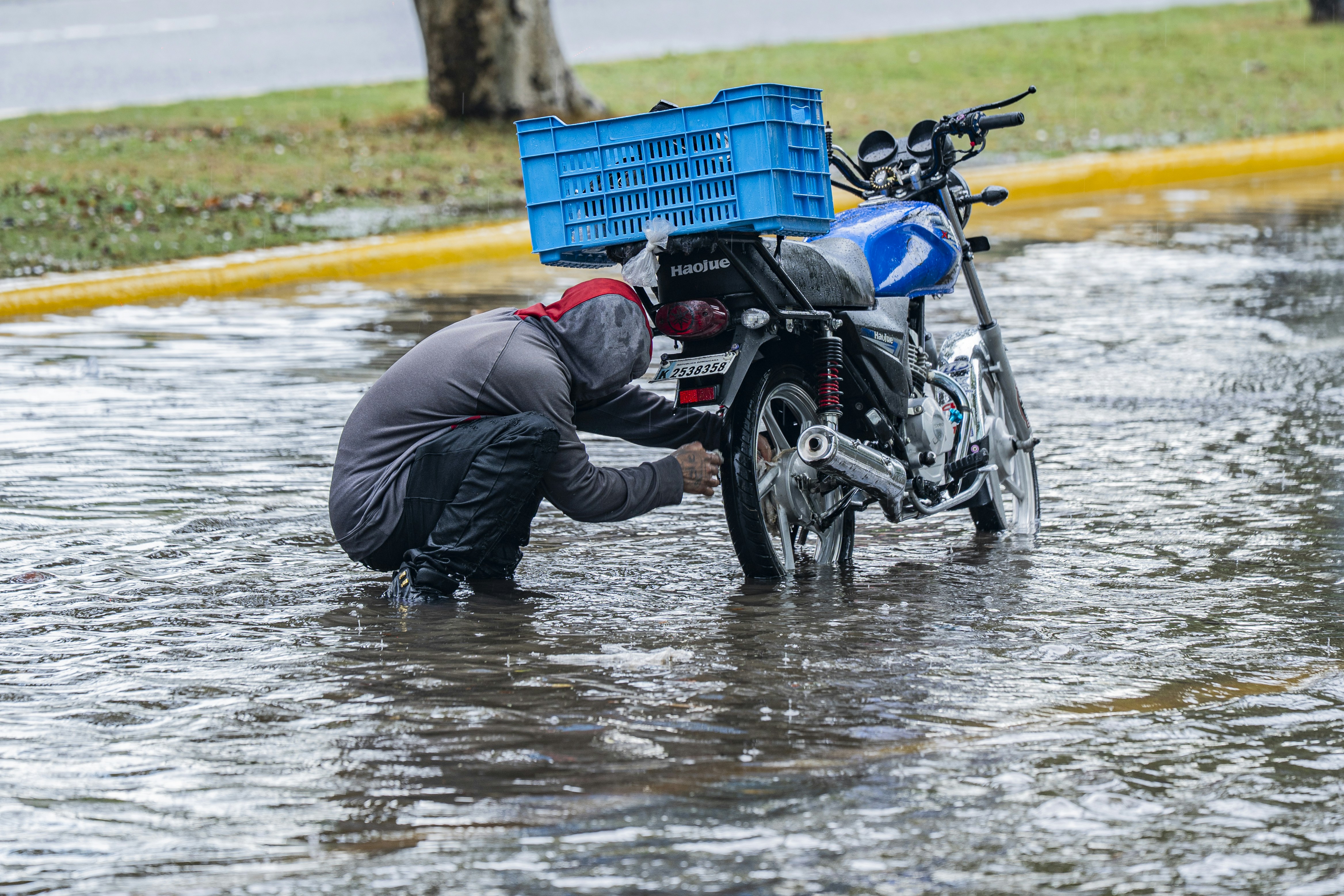 A person in a hooded sweatshirt crouching to fix a motorcycle tire in floodwater