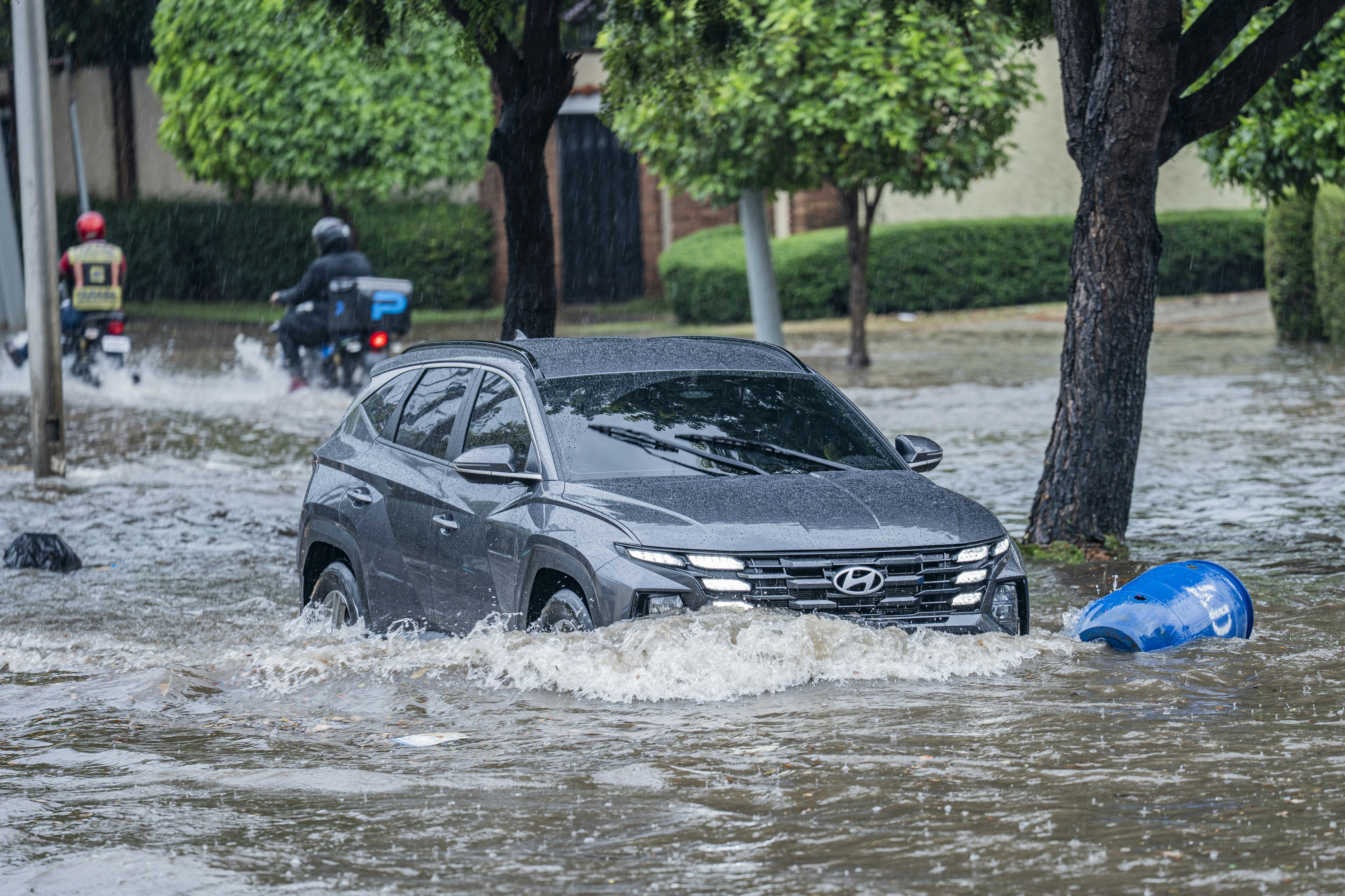 A grey SUV driving through a flooded street with motorcycles in the rain
